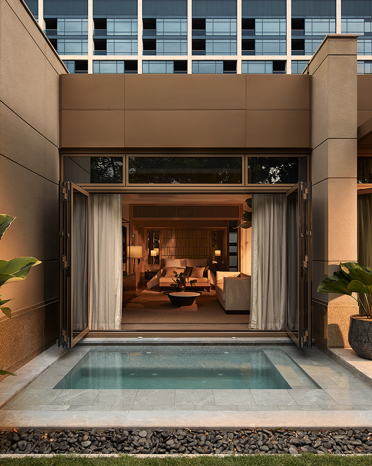 Elegant modern hotel exterior with an open living room visible through large glass doors. A small pool is in the foreground, surrounded by lush plants.