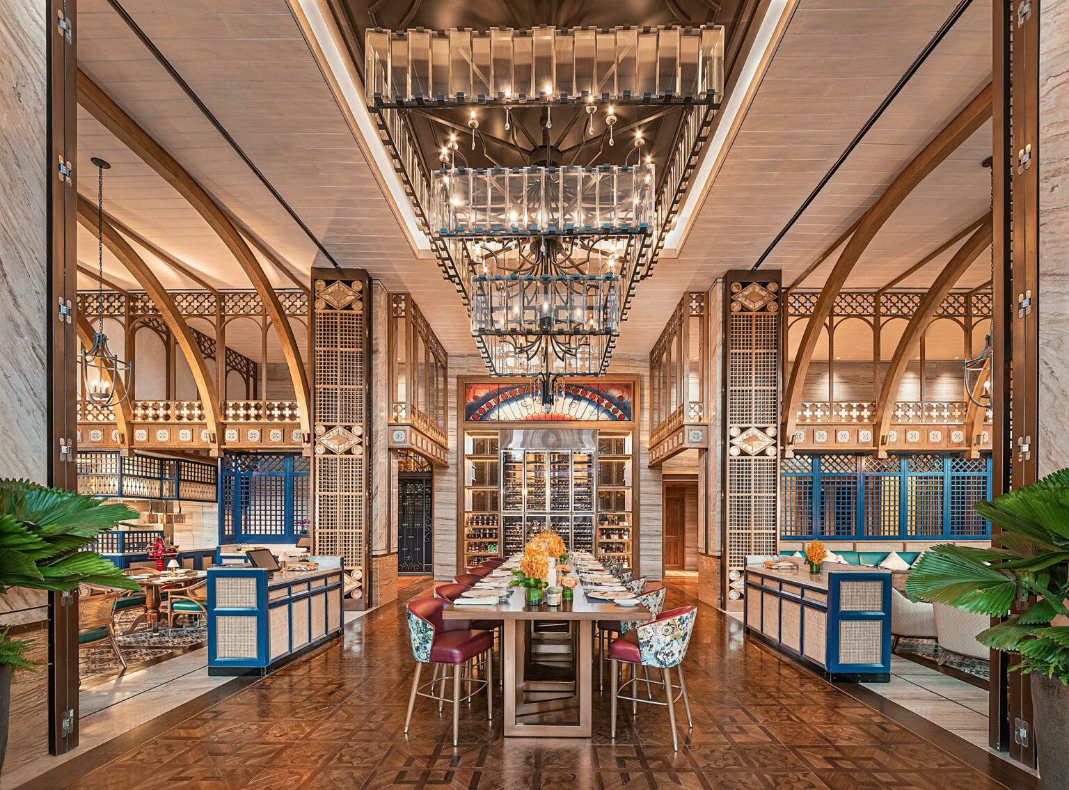 Elegant restaurant interior with ornate arches, large chandeliers, and a central table set for dining. Vibrant furnishings and symmetrical design create a luxurious ambiance.