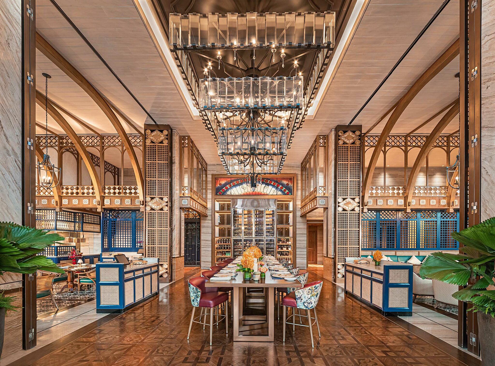 Elegant restaurant interior with ornate arches, large chandeliers, and a central table set for dining. Vibrant furnishings and symmetrical design create a luxurious ambiance.