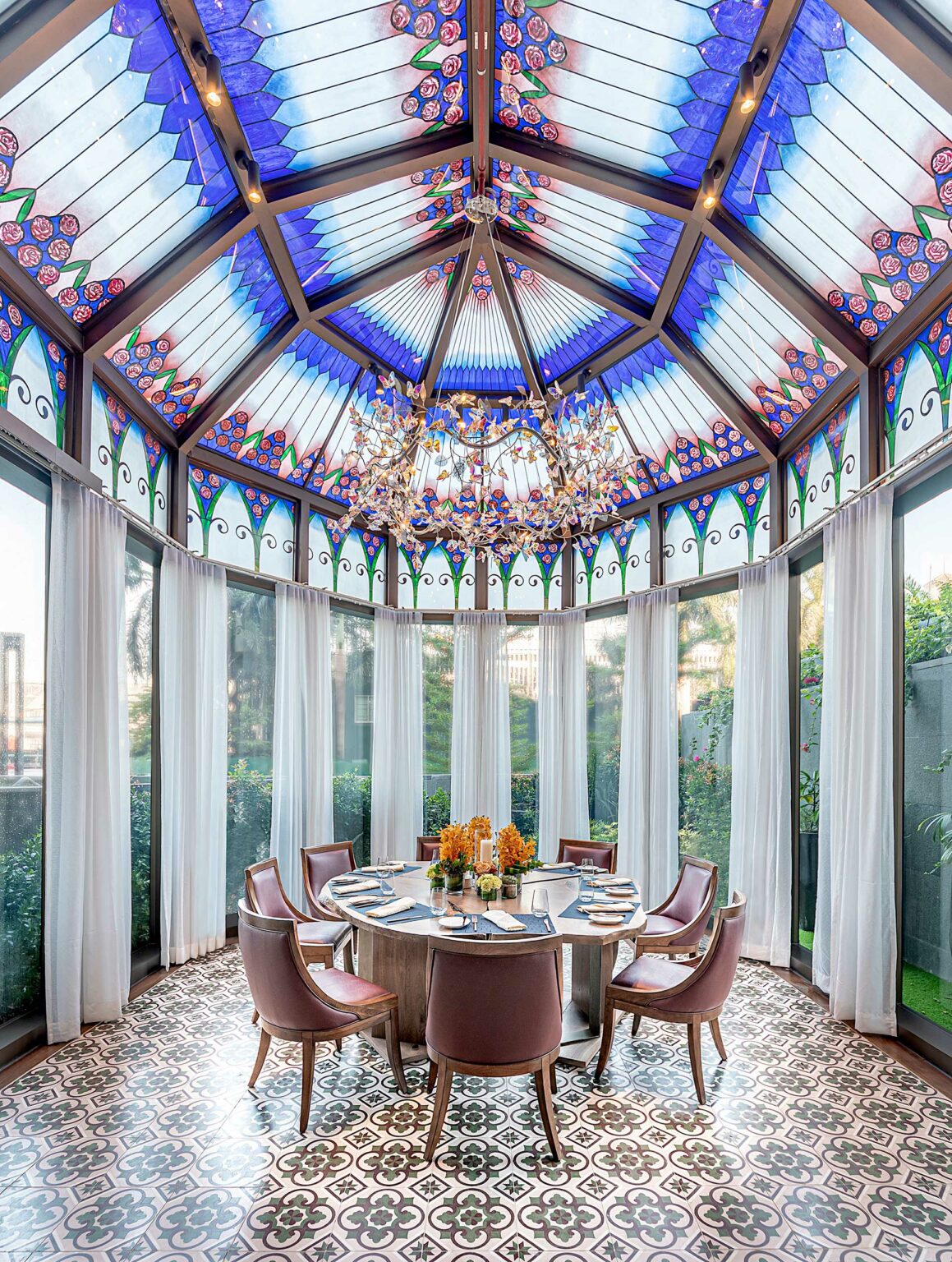 A private dining room of a restaurant with a round table and six leather chairs. The ceiling features vibrant stained glass in blue and purple. Sunlight streams through large windows with sheer curtains, creating an elegant and inviting atmosphere.