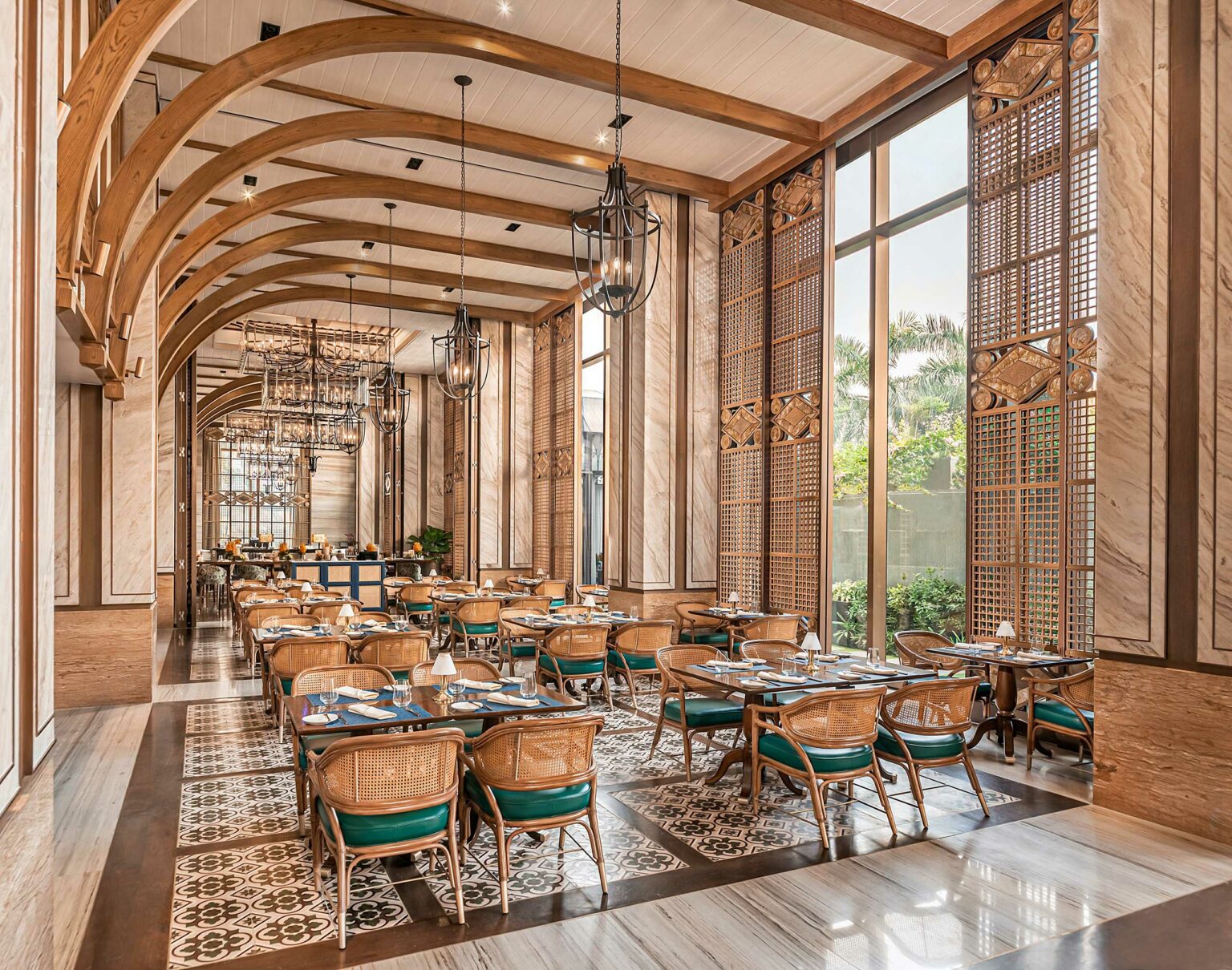 Elegant restaurant interior with high wooden arches, wicker chairs, and set tables. Large windows frame garden views, creating a warm, inviting ambiance.