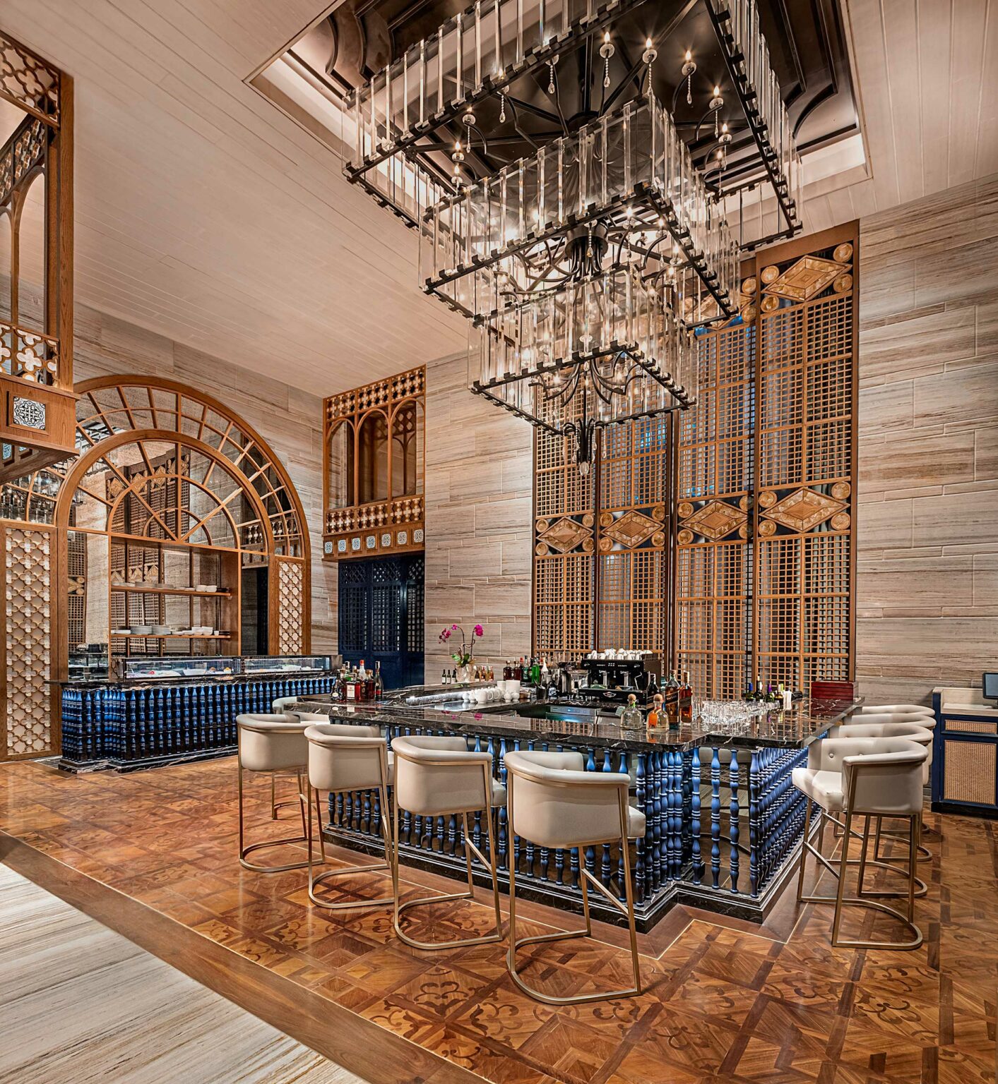 Luxurious bar area of a restaurant with a modern chandelier above a glossy black countertop. Elegant white chairs, intricate wooden lattice, and warm lighting create a sophisticated ambiance.