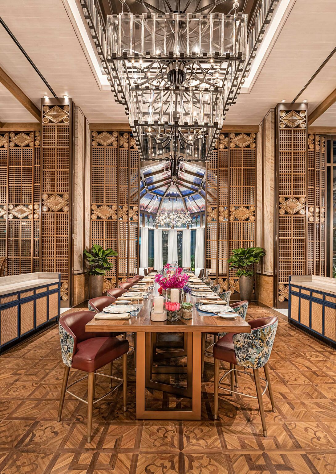 Elegant restaurant's dining room with a long wooden table set for a meal. Floral chairs, intricate wood walls, a large modern chandelier, and vibrant pink flowers create a luxurious atmosphere.