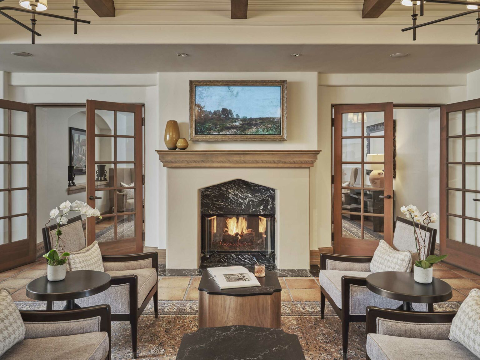 Cozy hotel lounge with a lit fireplace framed by marble, flanked by glass doors. Two armchairs, orchids, and soft lighting create an inviting atmosphere.