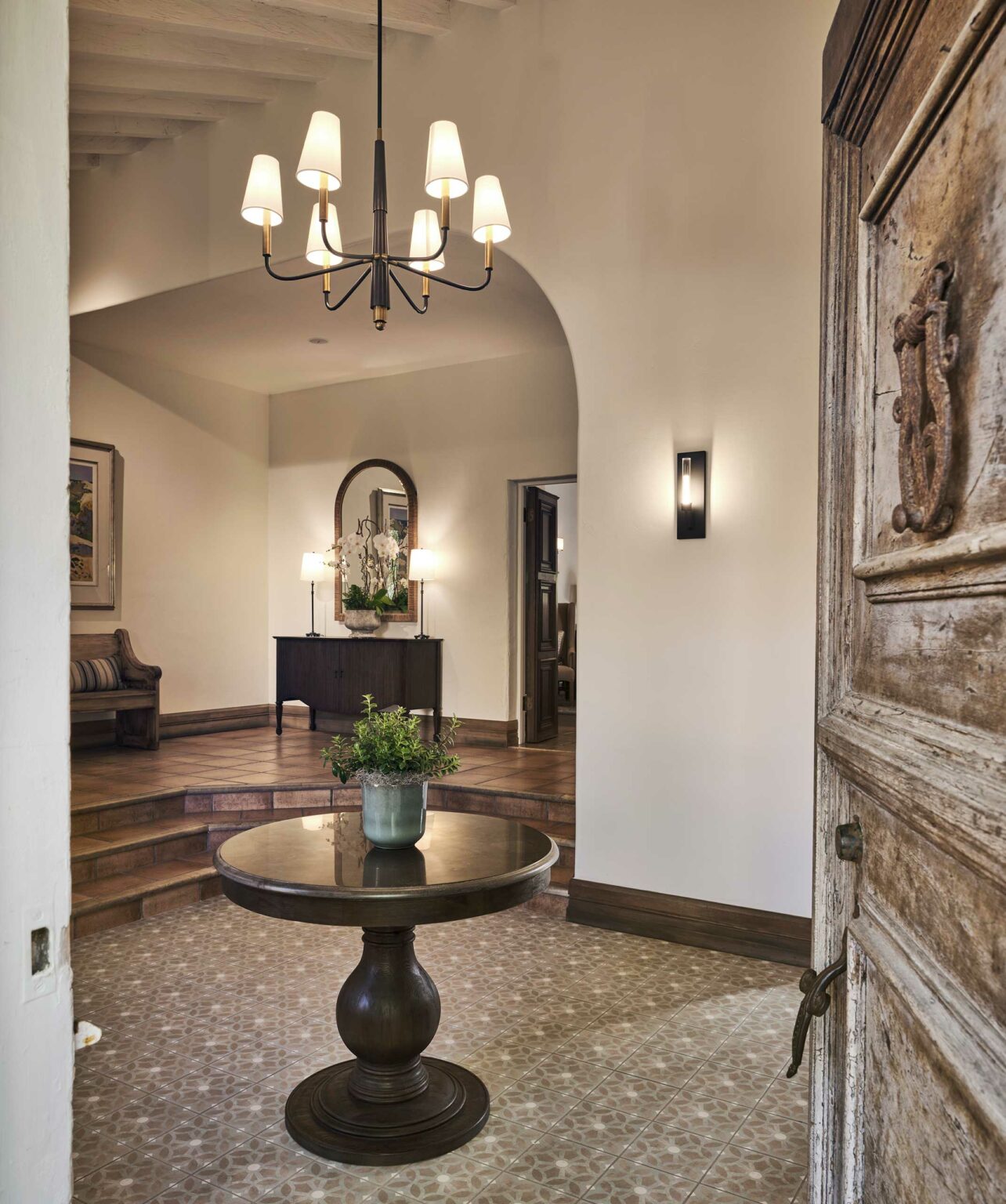 Warm, elegant entryway featuring a round wooden table with a potted plant, rustic door, chandelier, and arched doorway with a console and mirror in view.