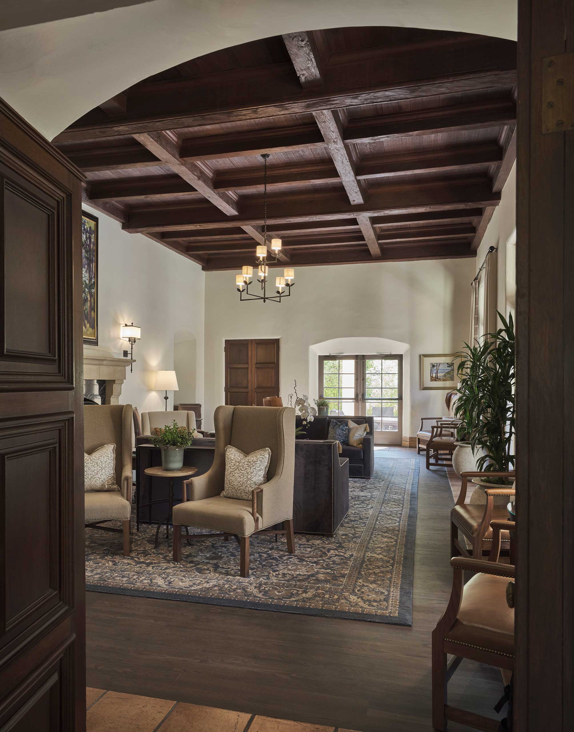 Elegant suite living room with high, dark wood-beamed ceiling, plush armchairs, and a large patterned rug. Natural light streams through a window, creating a warm, inviting atmosphere.