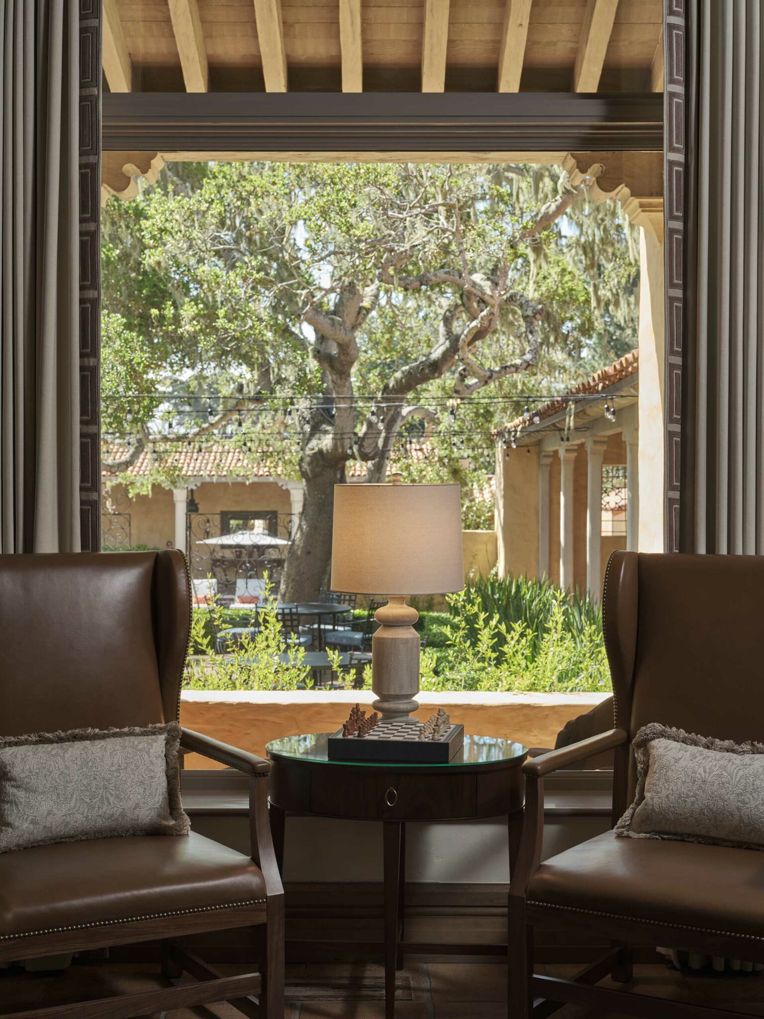 Suite living room scene, featuring two leather chairs with cushions face a round table with a lamp. Beyond, a window reveals a tree and courtyard with rustic charm. Warm and inviting ambiance.