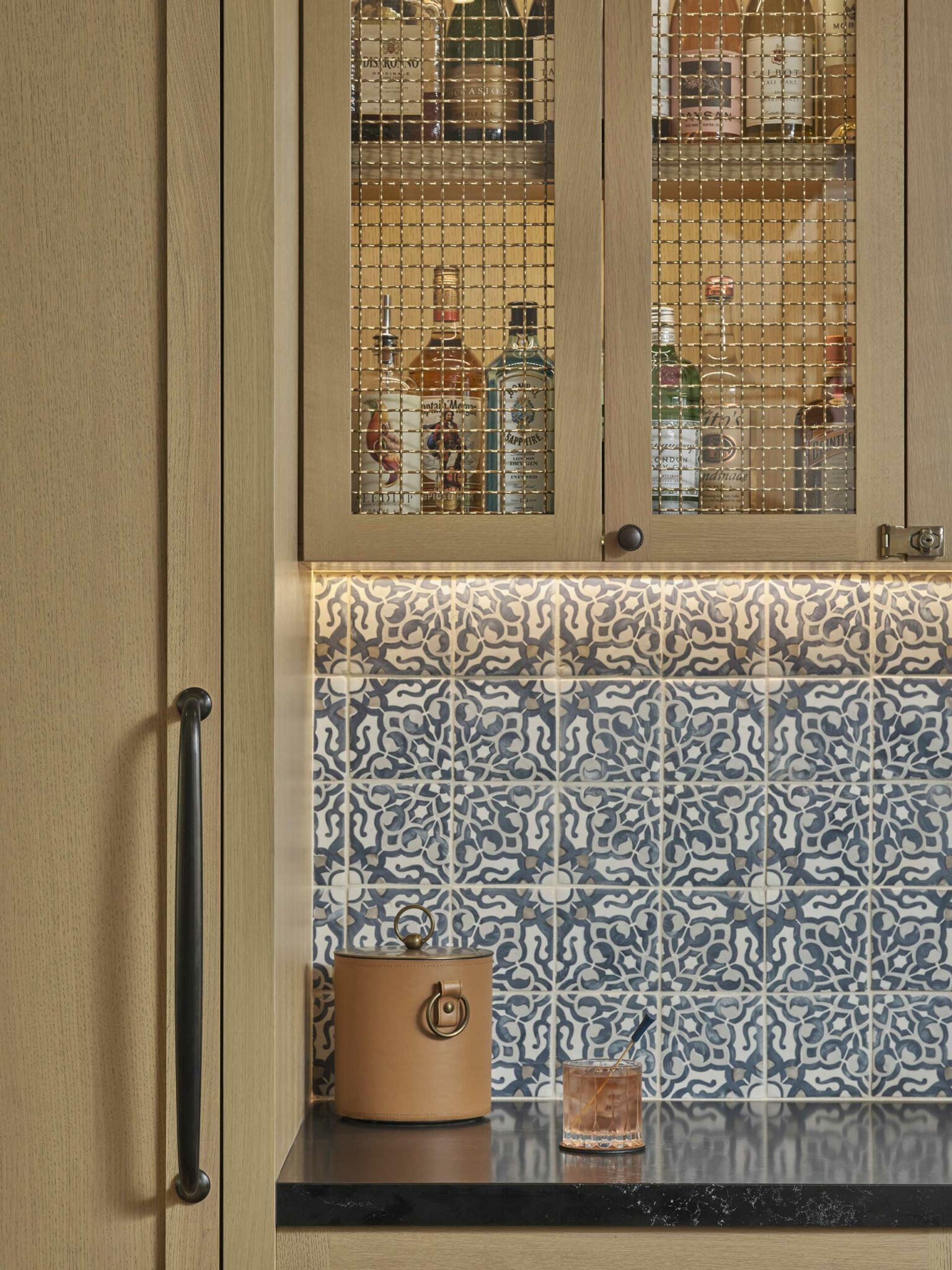 A cabinet with a wire-mesh door shows liquor bottles inside. Below, blue patterned tiles and a countertop hold a brown container and a glass with amber liquid.