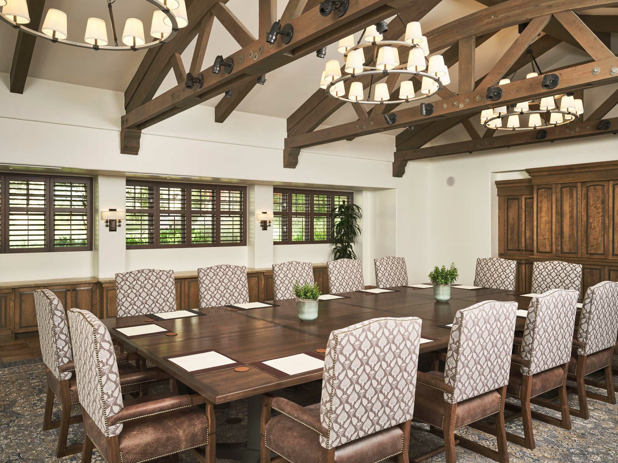 Spacious hotel conference room with a large wooden table surrounded by patterned chairs. Natural light filters through shuttered windows. Elegant chandeliers hang from exposed beams.