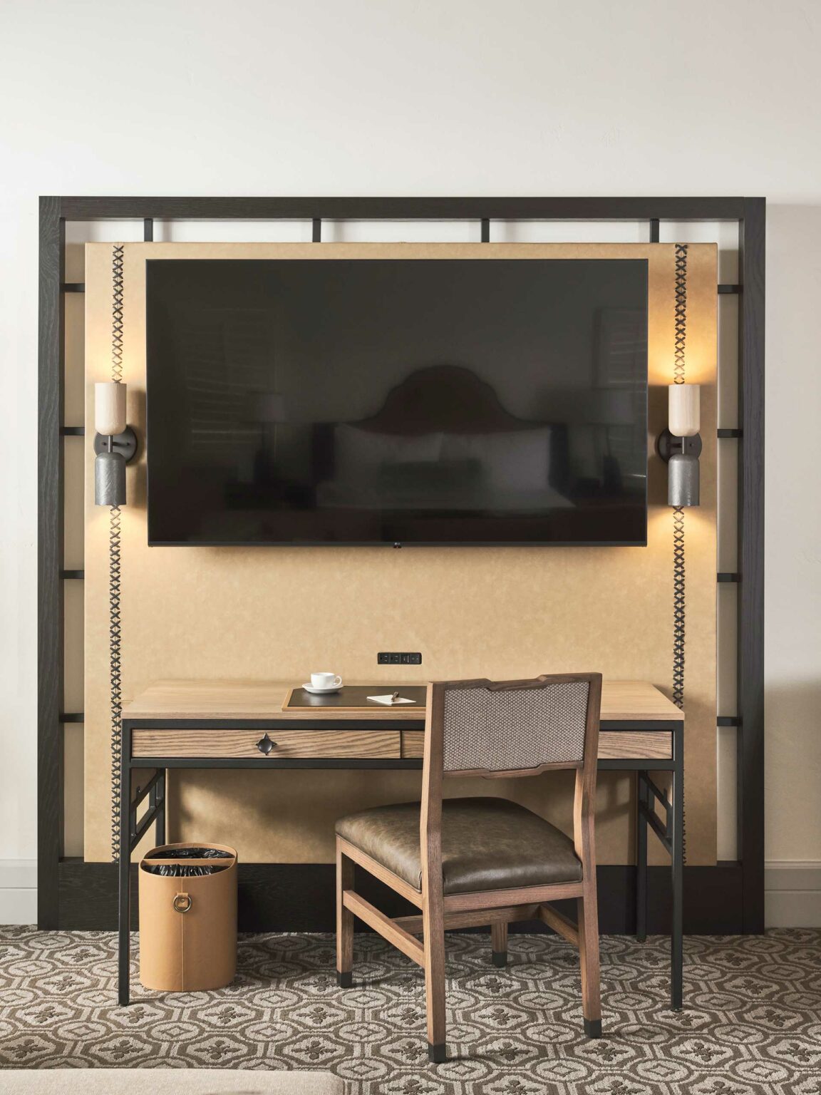 Stylish hotel room with a large flat-screen TV on the wall above a wooden desk. A cushioned chair faces the desk, and a waste bin is nearby.