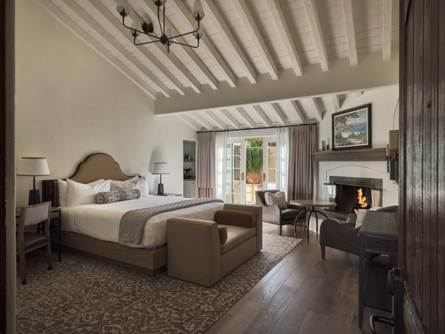 Elegant suite bedroom with vaulted ceiling, cozy fireplace, and plush king-size bed. French doors open to a patio, enhancing the warm and inviting ambiance.