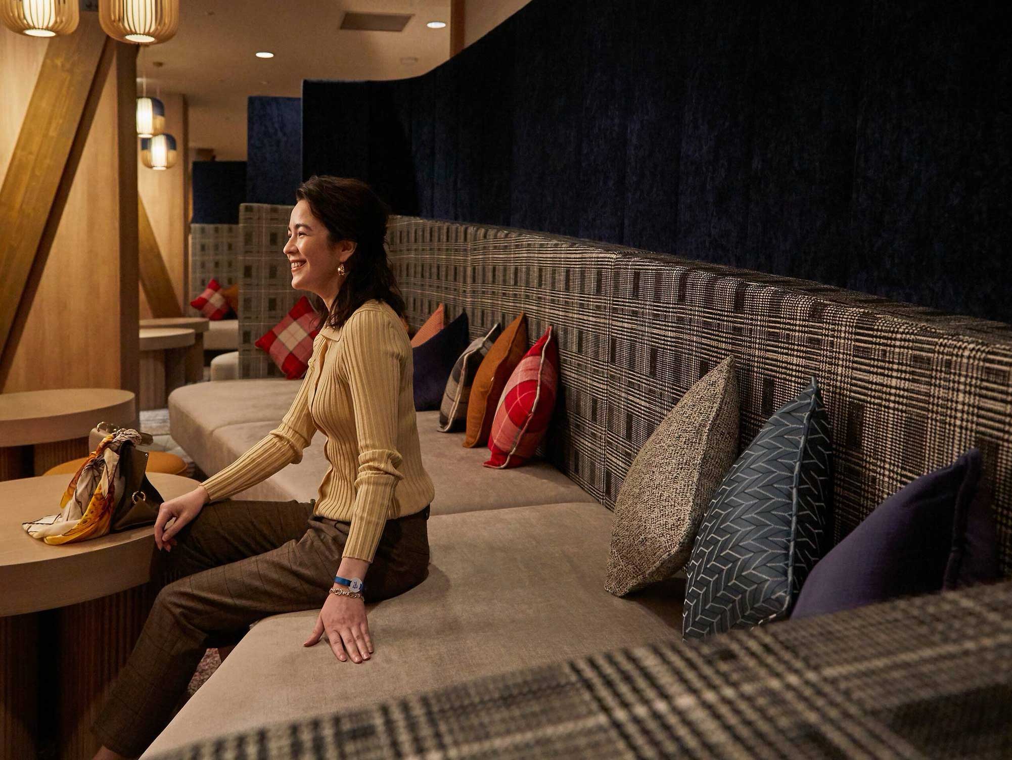 A woman in a beige sweater sits smiling on a cushioned bench with colorful pillows in a cozy, warmly lit hotel lounge. The atmosphere is relaxed and inviting.