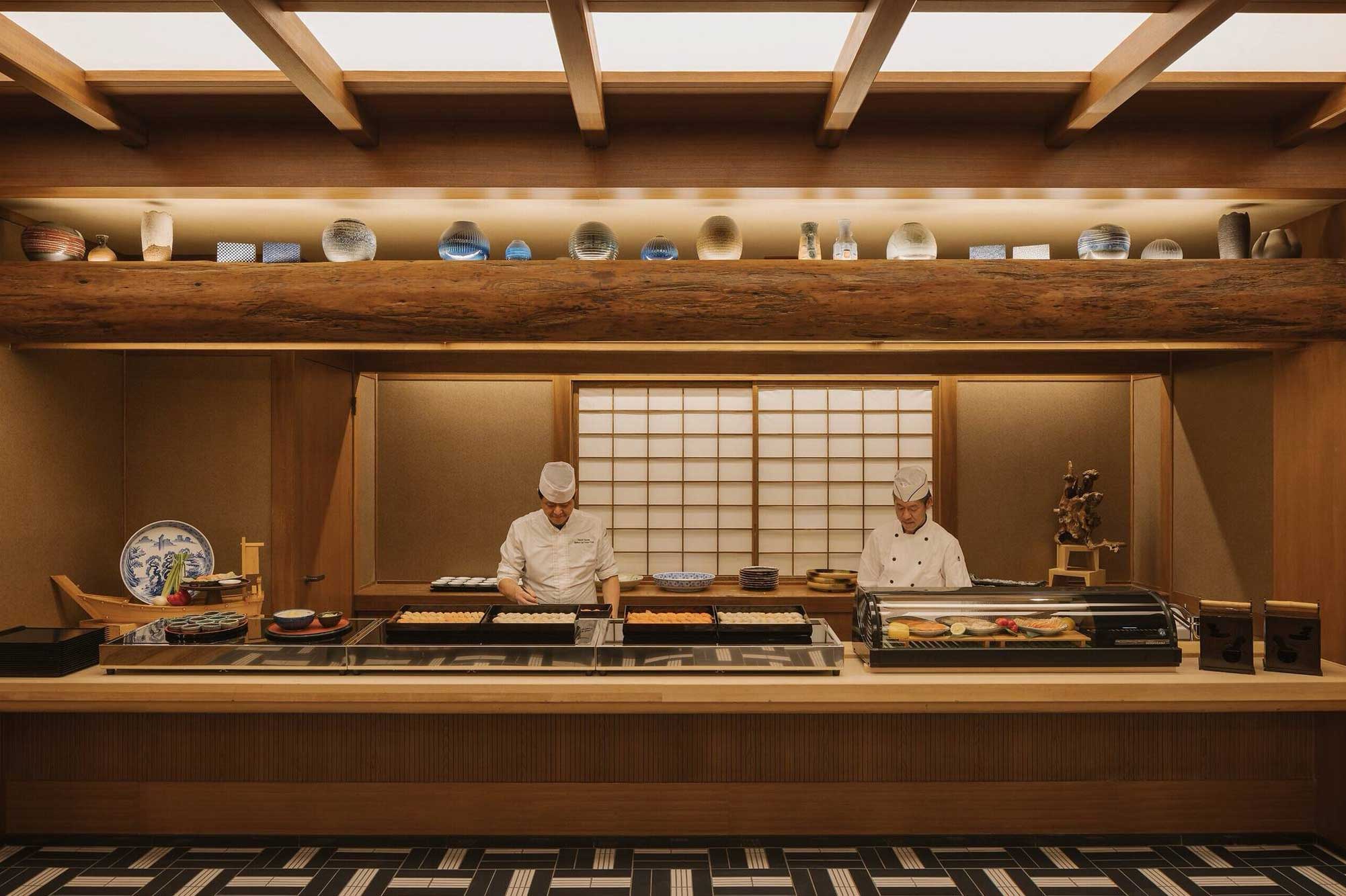 Two chefs in white uniforms prepare food behind a Japanese-style wooden counter. Shelves with decorative plates and bowls are above, creating a calm and elegant ambiance.