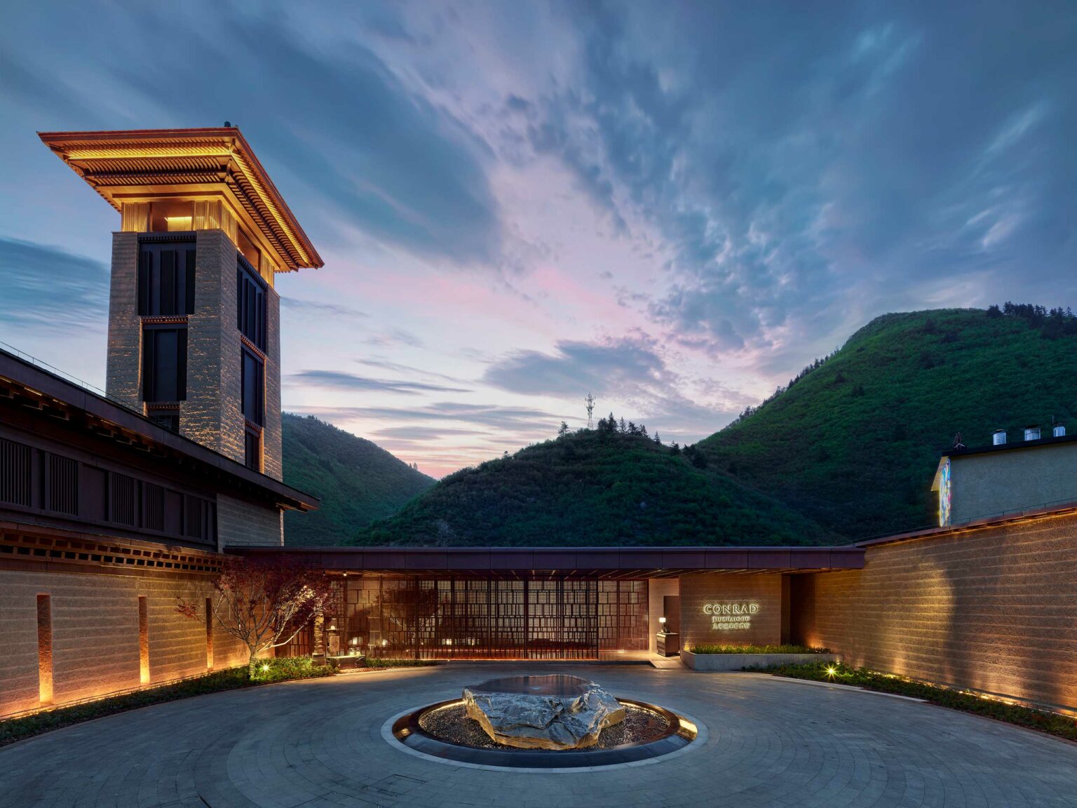 Modern hotel building with a tall tower and courtyard at dusk, surrounded by green hills under a vibrant sky. Warm lights create a tranquil ambiance.