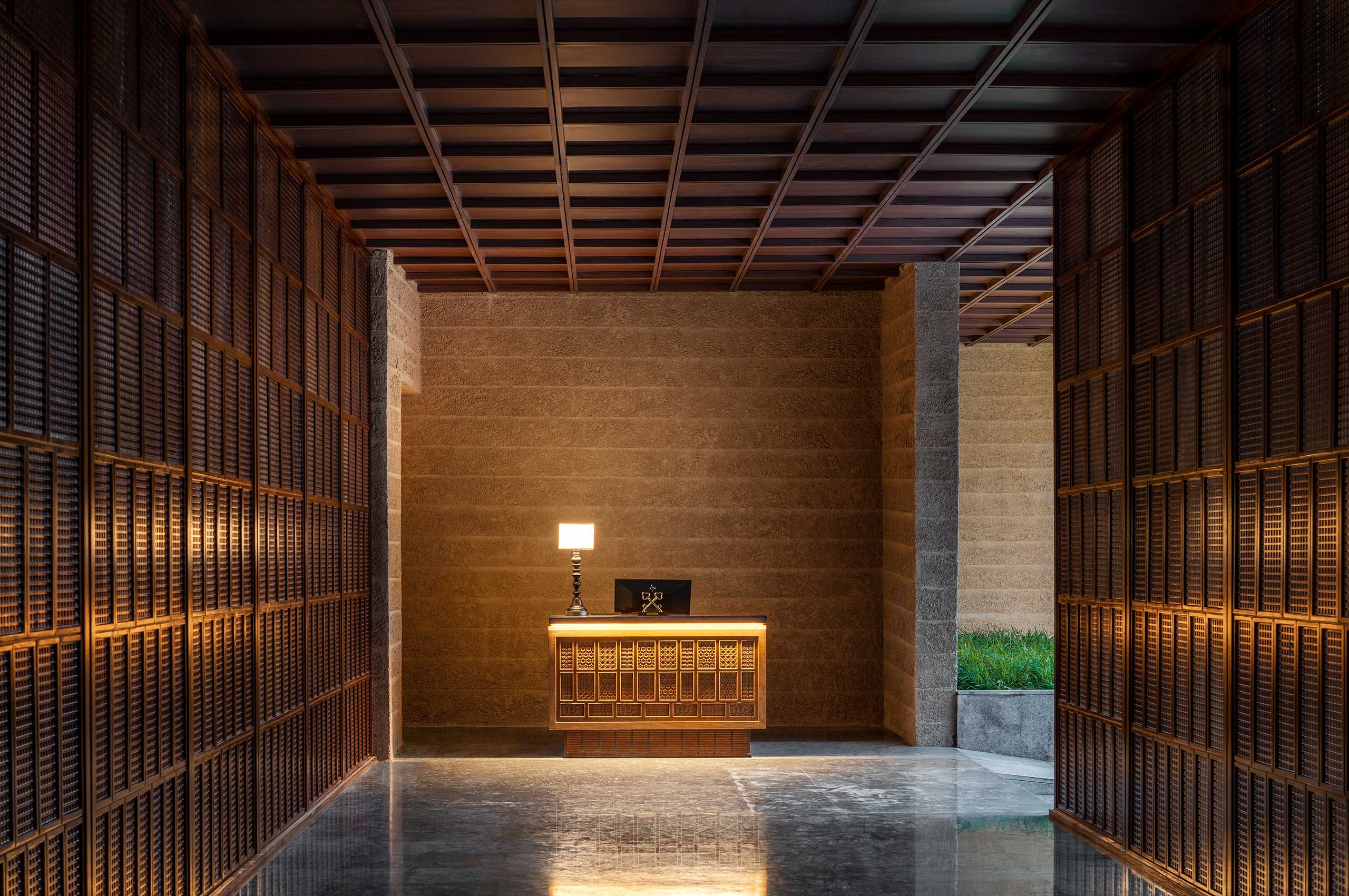 Hotel reception area with warm lighting, featuring a wooden desk and table lamp against a textured wall. Flanked by lattice-patterned panels, creating an inviting atmosphere.