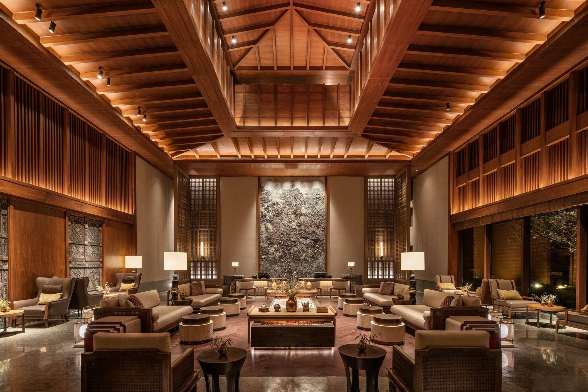 Luxurious hotel lobby with high wooden ceilings, plush seating, and warm lighting. A large, textured wall art centerpiece adds elegance. Cozy and inviting.