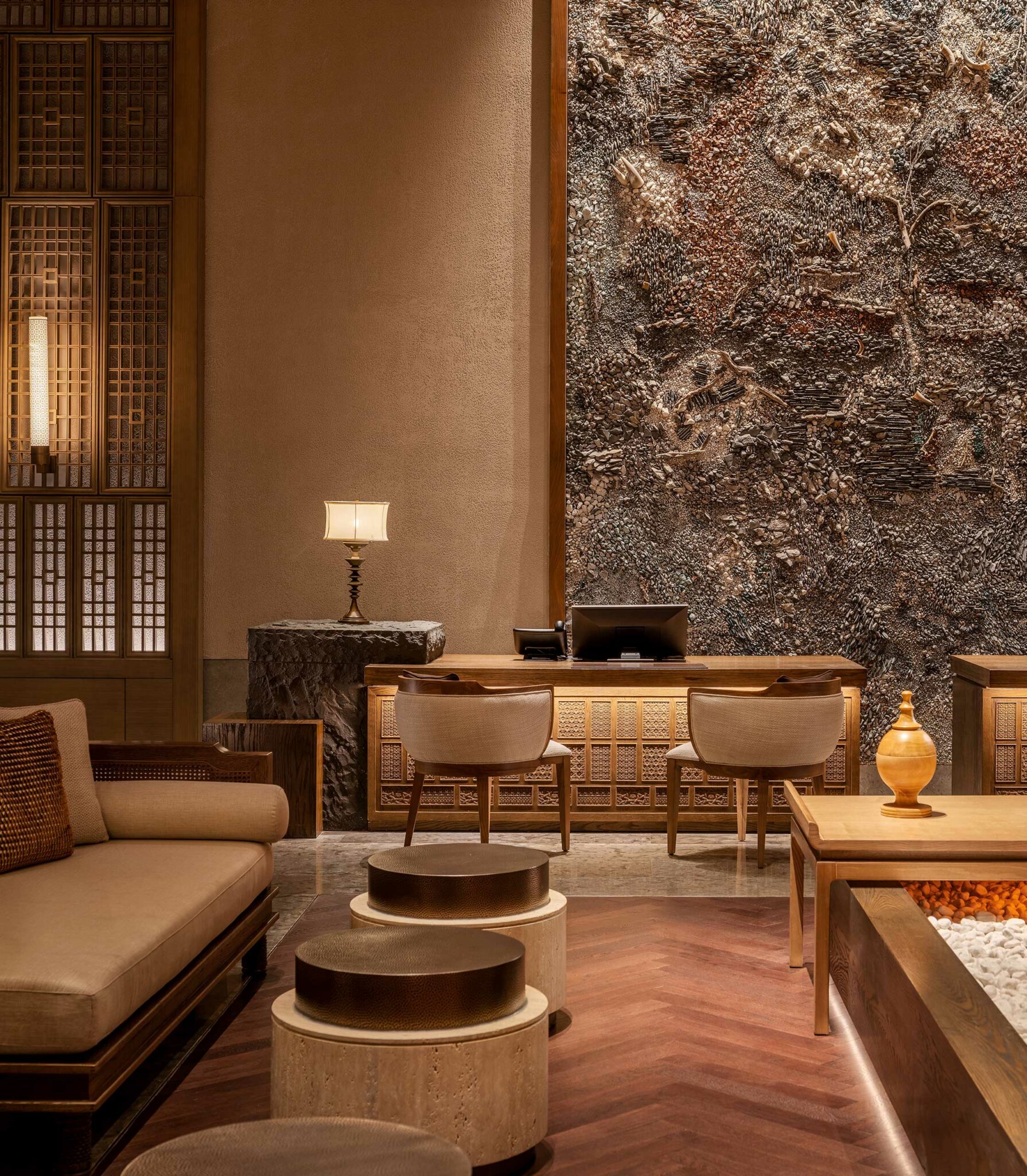 Luxurious hotel lobby with a warm, earthy tone, featuring a textured stone wall, elegant seating, a wooden desk, soft lighting, and natural decor.