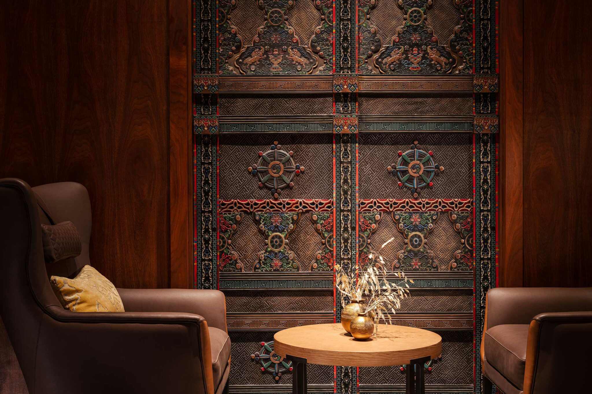 Elegant hotel lounge nook with two brown leather chairs and a wooden table in between. Ornate, intricate wall paneling in the background adds a luxurious feel.