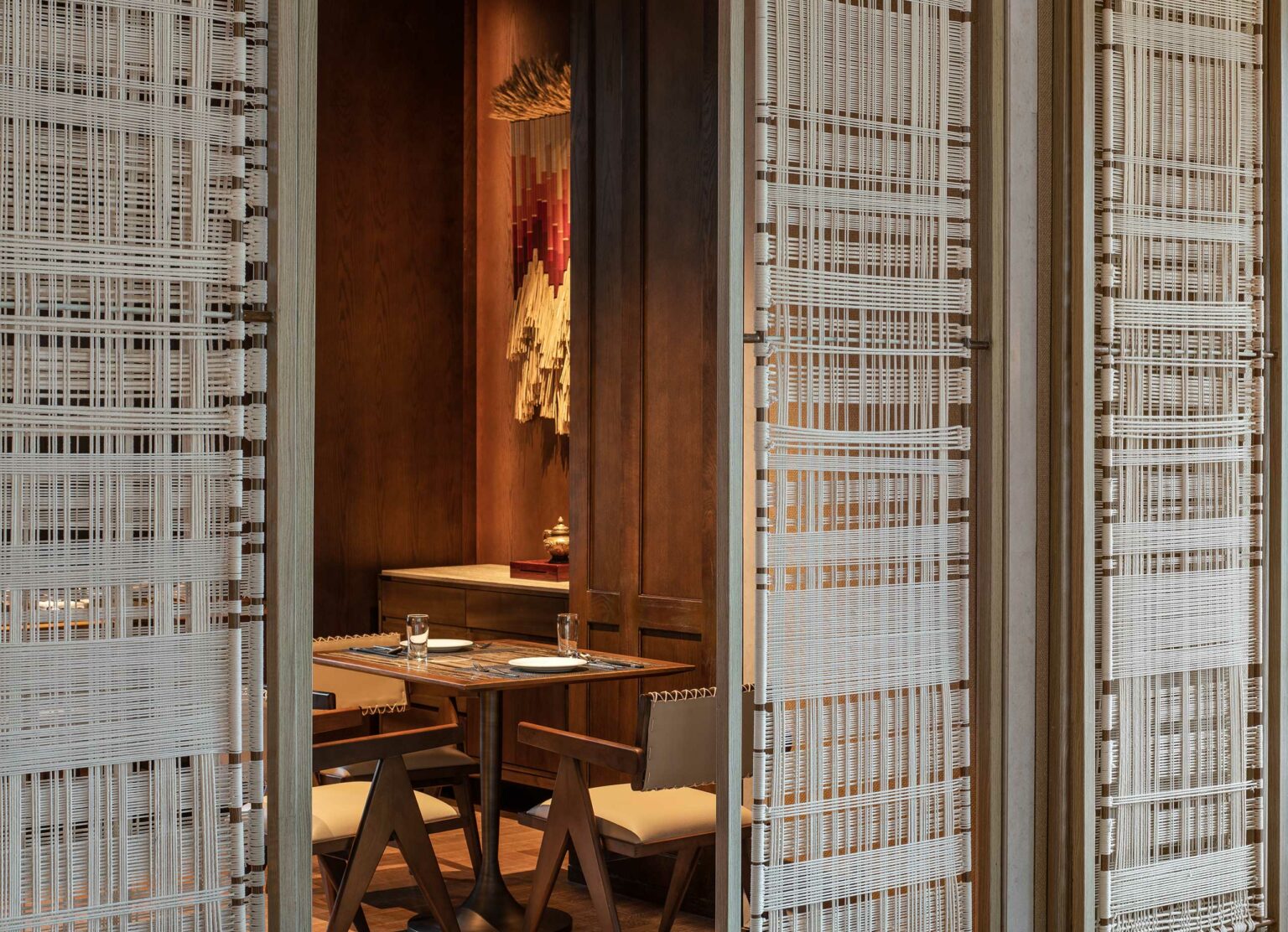 Cozy hotel dining nook with a wooden table set for two, surrounded by woven partitions. A wall hanging adds a warm, inviting touch to the elegant décor.