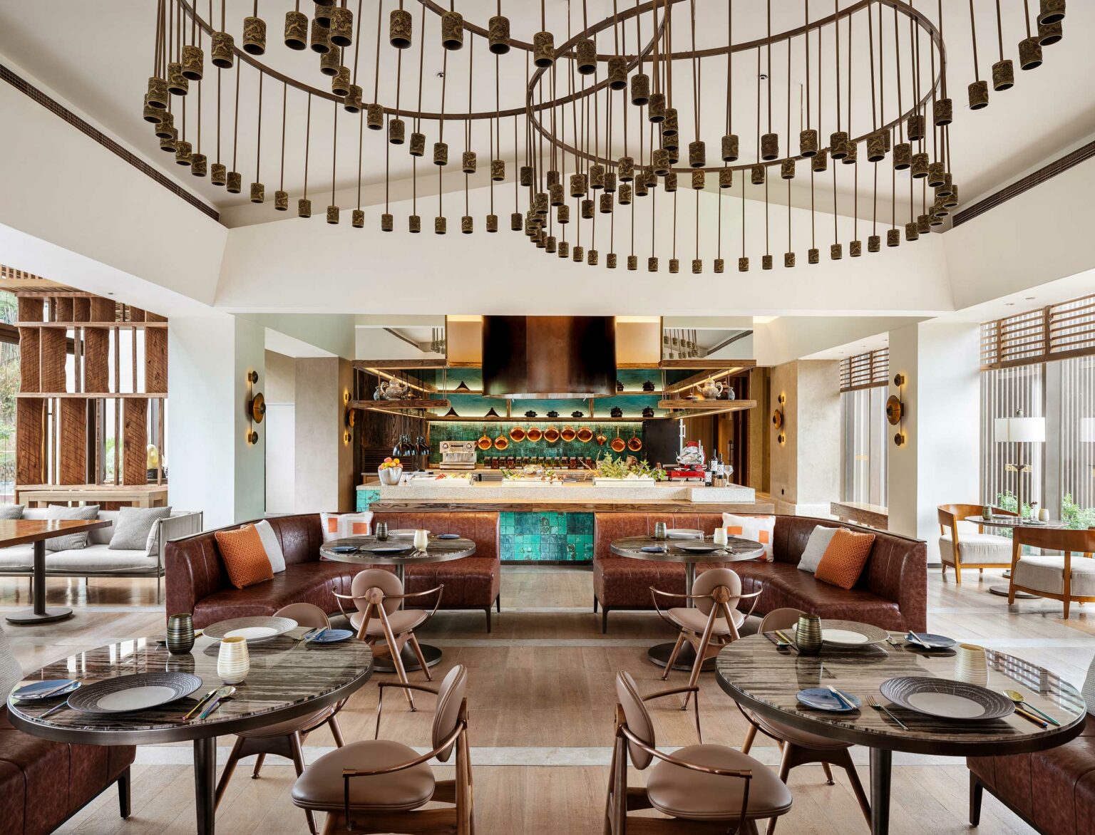 Modern hotel all day dining with circular tables, brown leather couches, and a decorative ceiling light. The open kitchen displays copper pots. Cozy and inviting ambiance.