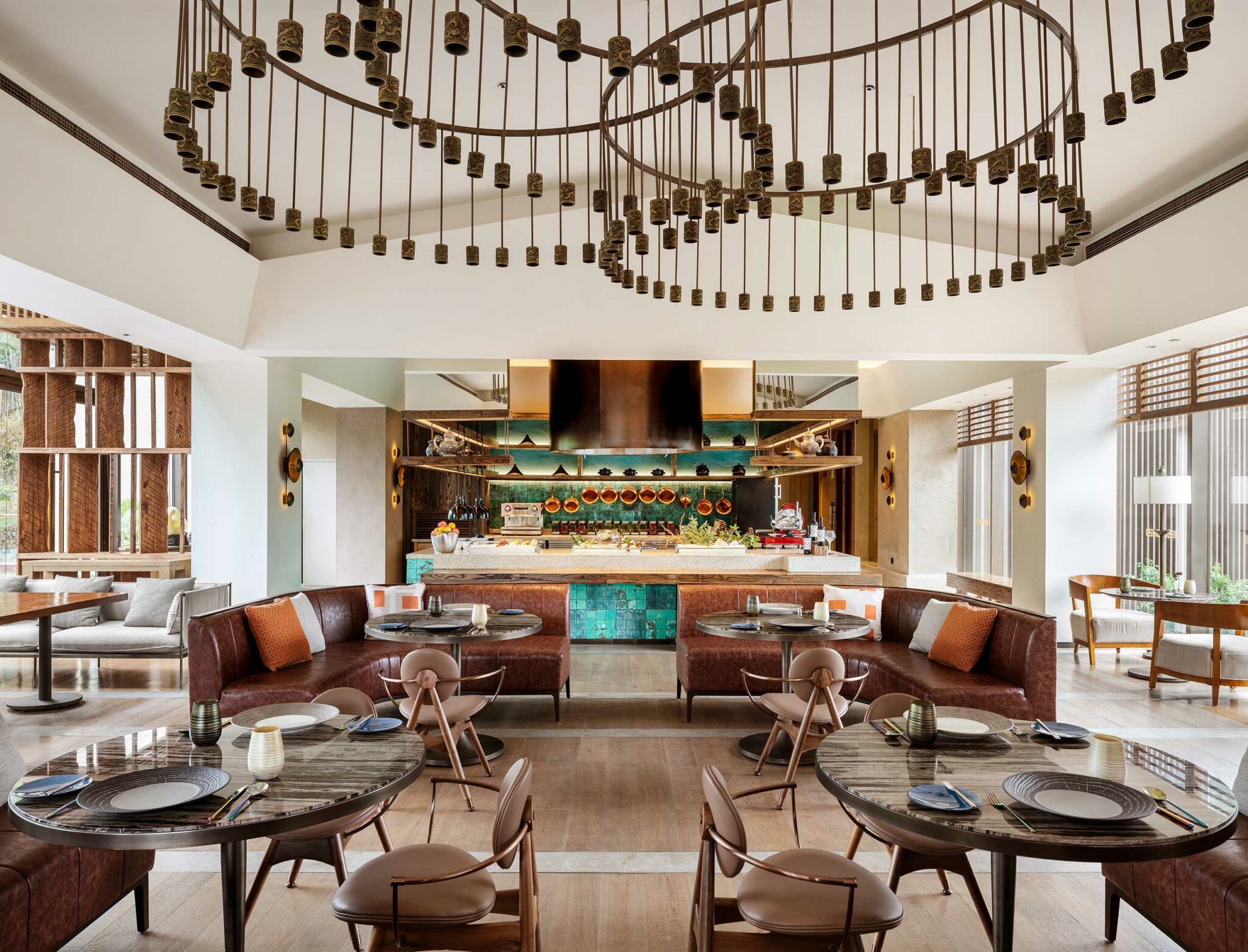 Modern hotel all day dining with circular tables, brown leather couches, and a decorative ceiling light. The open kitchen displays copper pots. Cozy and inviting ambiance.