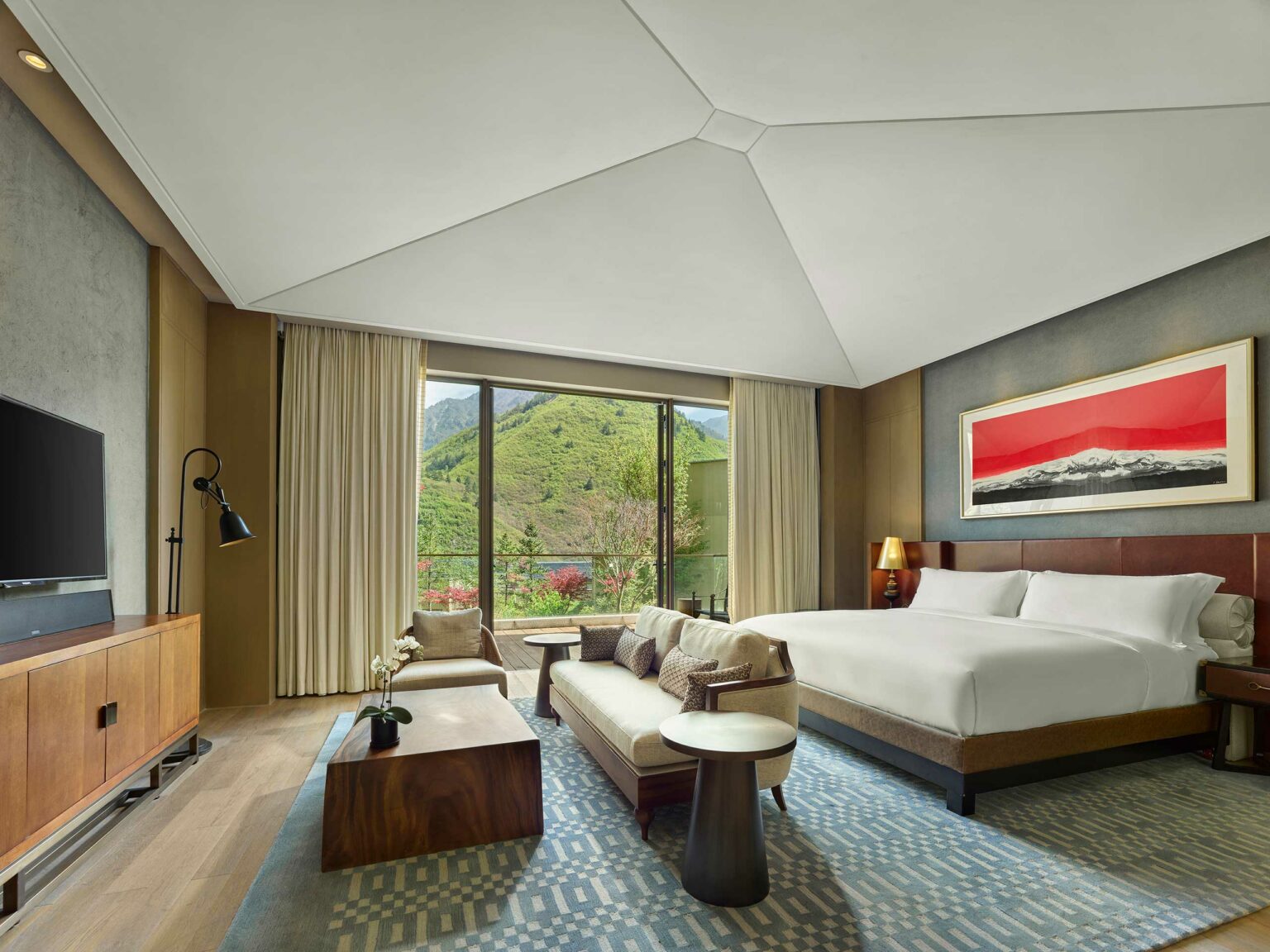 Modern hotel room with a large bed and seating area facing glass doors revealing a scenic mountain view. The atmosphere is serene and inviting.