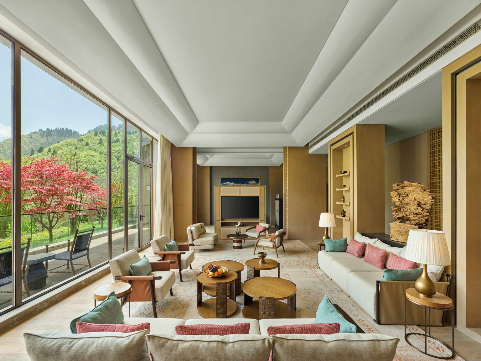 Spacious, elegant hotel lounge with modern beige sofas, colorful cushions, and wooden tables. Large windows reveal lush green hills and a red tree outside.