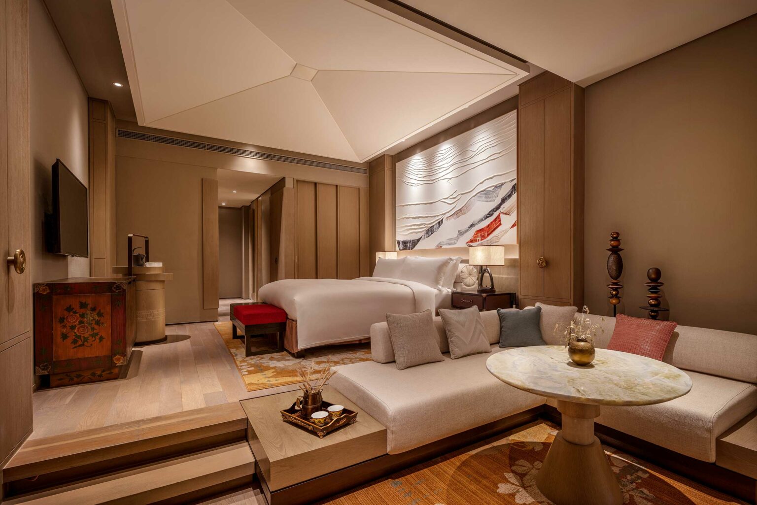 Elegant hotel room featuring a large bed with plush pillows, a cozy seating area with a round table, warm neutral tones, and artistic wall decor.