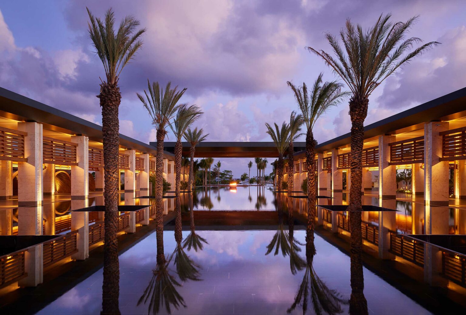 A serene resort courtyard with tall palm trees lining a reflective water pool, under a purple sky at sunset. Warm lights create a tranquil, luxurious ambiance.