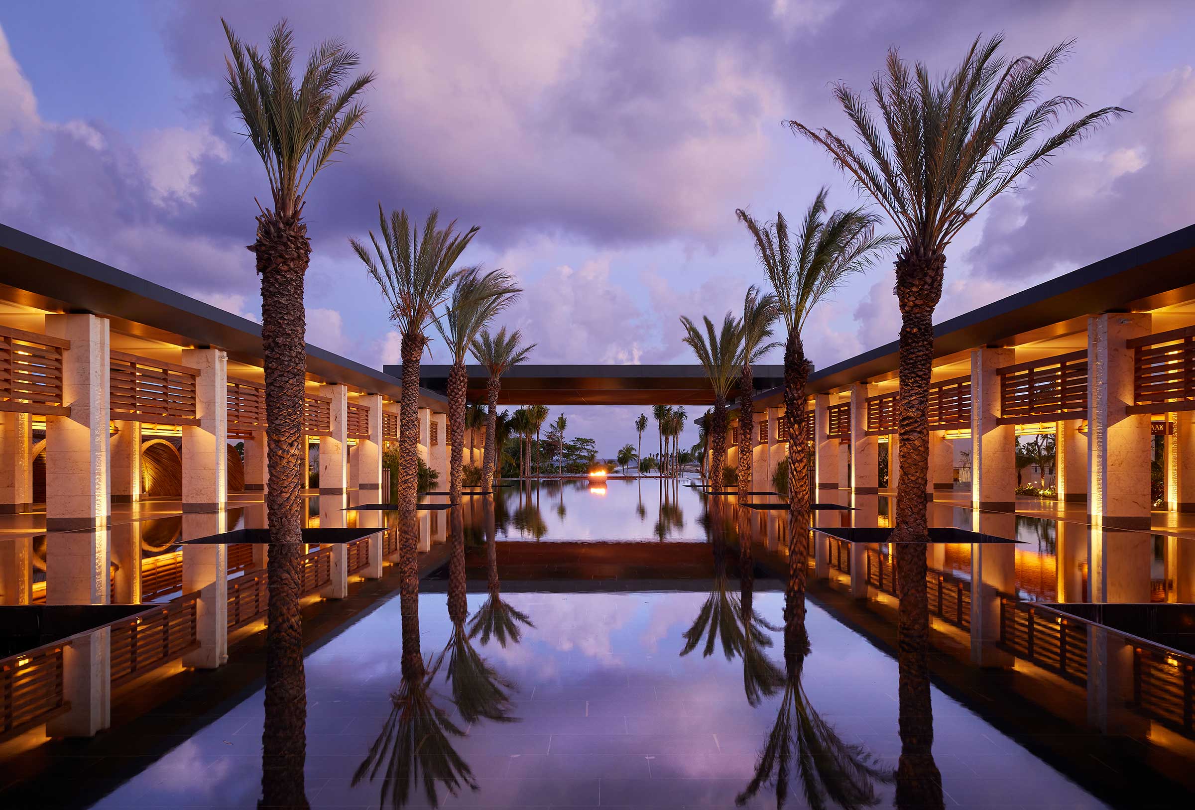 A serene resort courtyard with tall palm trees lining a reflective water pool, under a purple sky at sunset. Warm lights create a tranquil, luxurious ambiance.