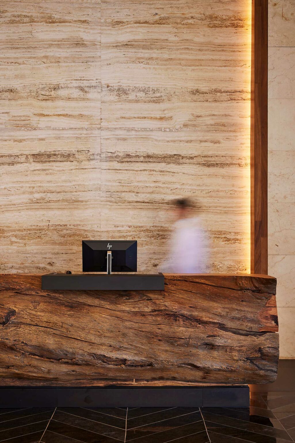 Resort reception area with a large, rustic wooden desk, a computer monitor on top, and a blurred figure walking by. The background features textured stone walls with soft lighting.