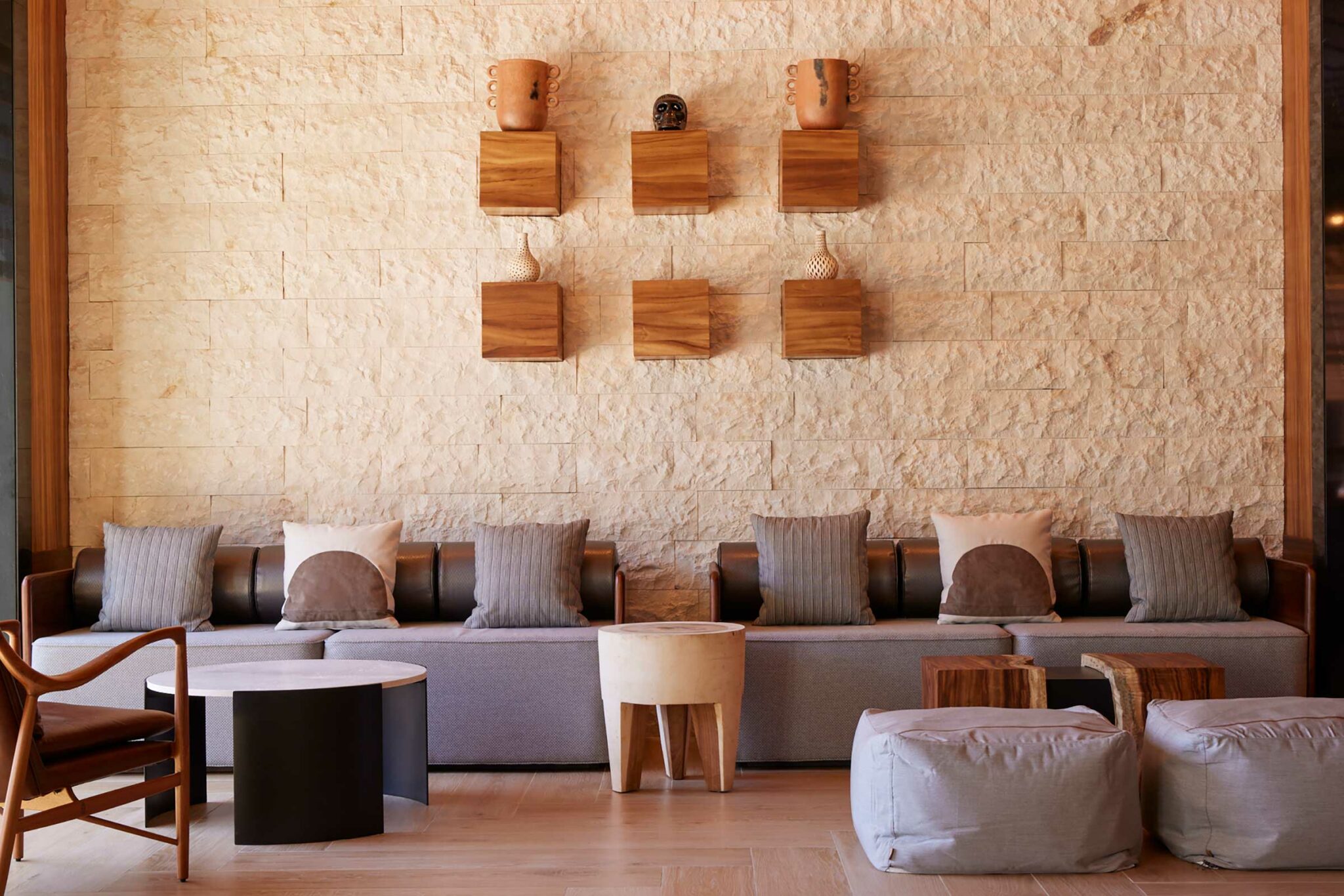 Modern resort lounge with a textured stone wall, featuring wooden shelves displaying pottery. A gray sofa with soft cushions, stylish tables, and poufs add warmth.