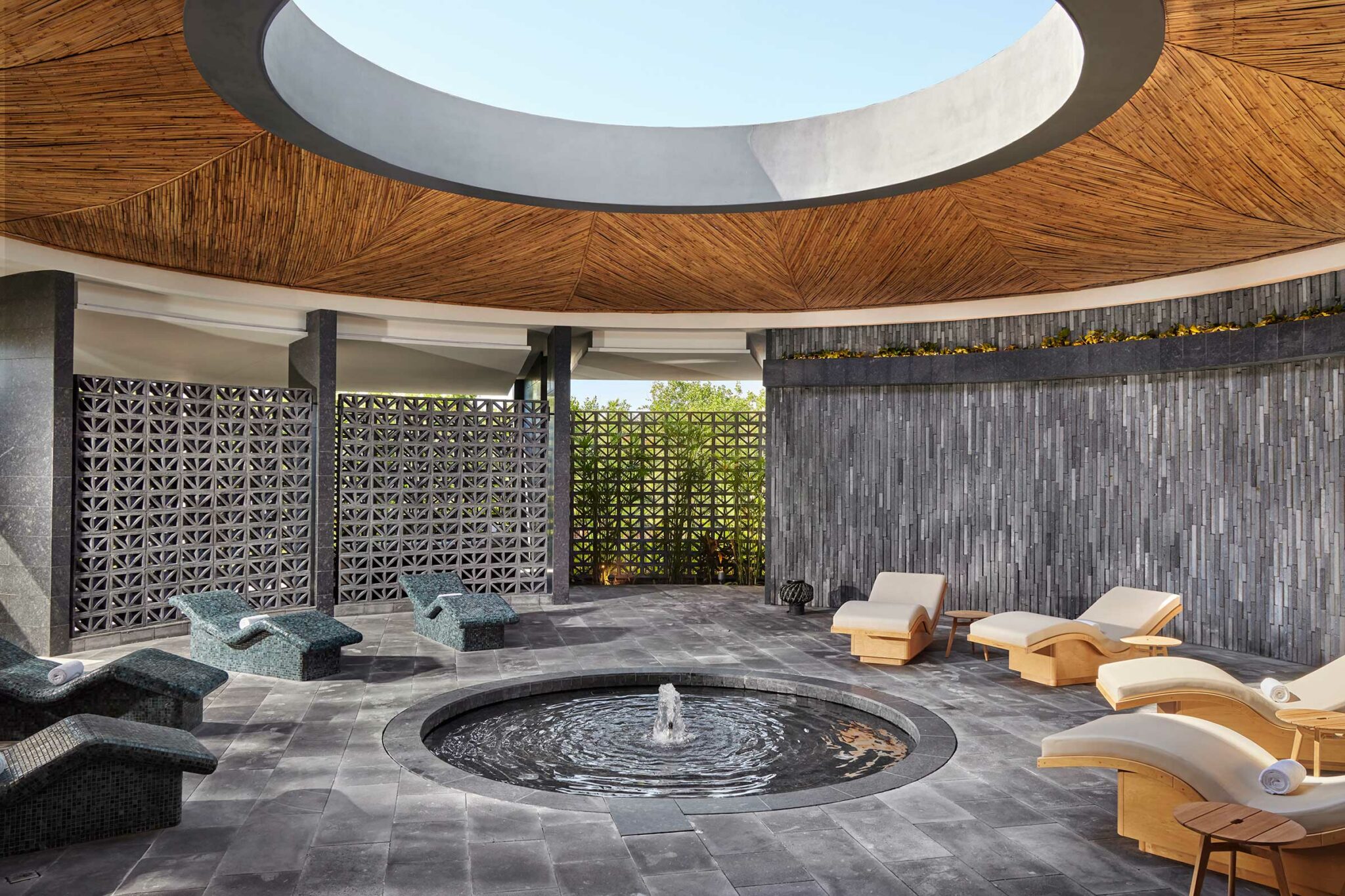 Spacious resort spa interior with a circular skylight, bamboo ceiling, modern loungers, and a central water feature. The ambiance is serene and luxurious.