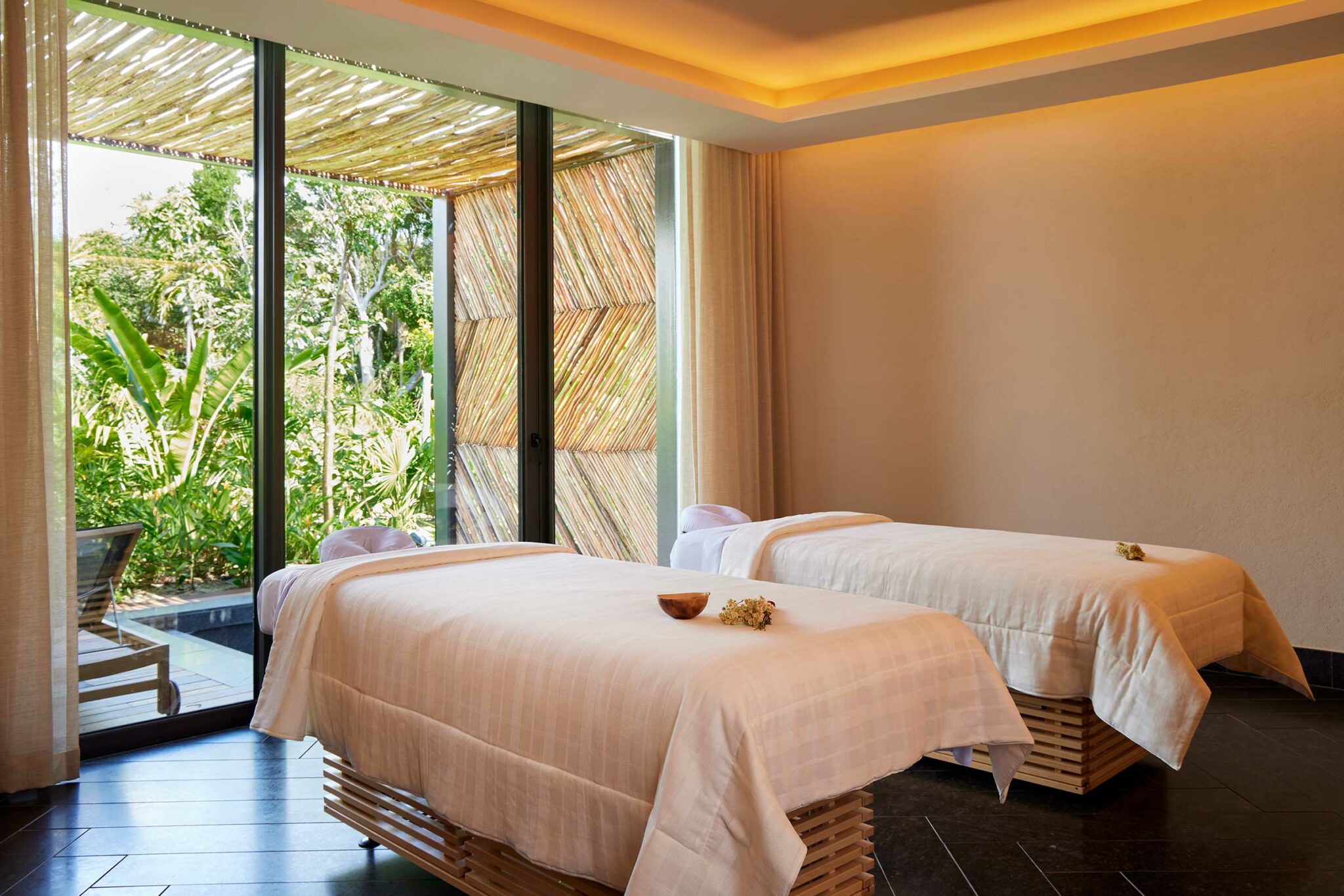 Two resort spa treatment beds with white linens in a serene room, overlooking lush greenery through large windows. Soft lighting creates a calming atmosphere.