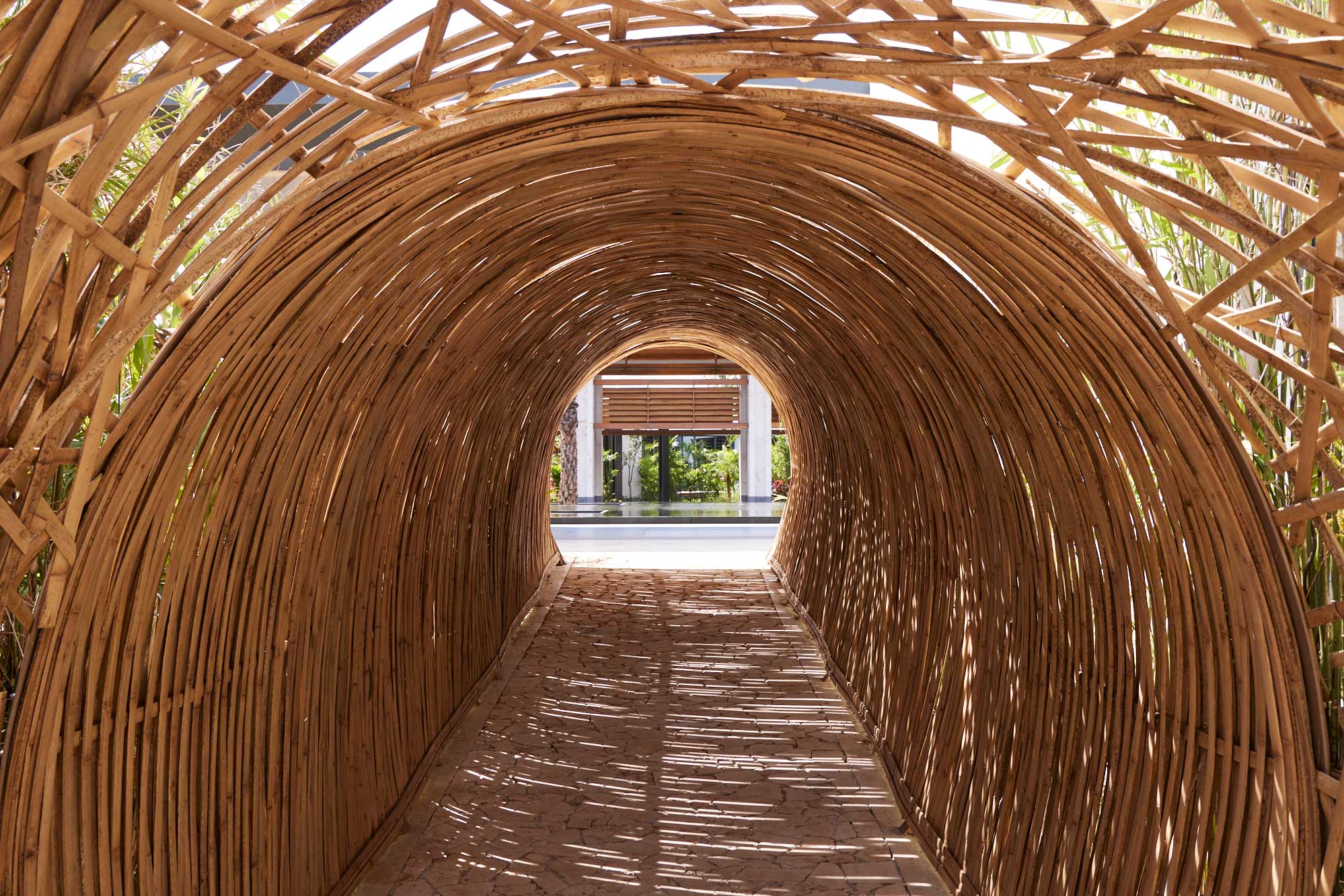 A tunnel made of interwoven bamboo slats, creating an arched resort pathway. Sunlight filters through, casting shadows on the ground, with greenery visible at the exit.