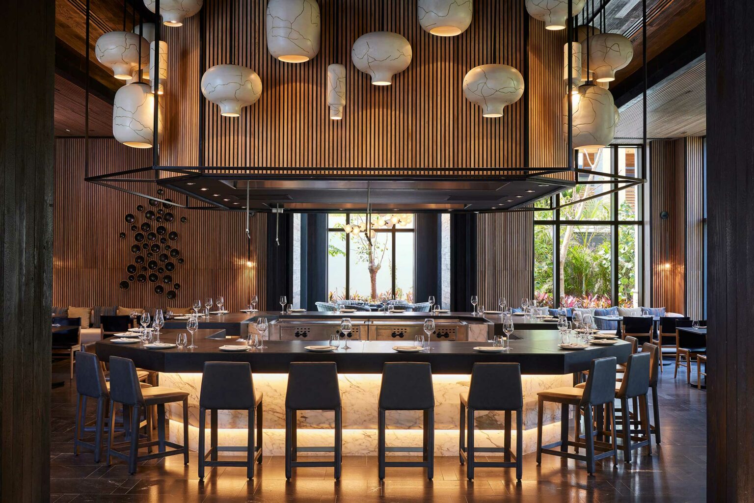 Modern resort restaurant interior with a circular bar, sleek grey chairs, and an illuminated marble counter. Hanging lamps and wooden panels enhance elegance.