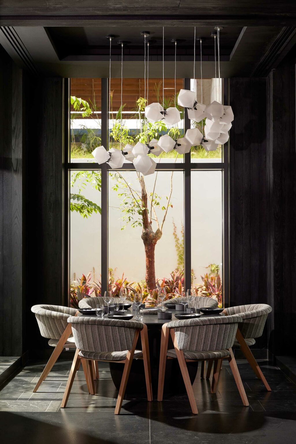 A cozy resort dining area with a round table set for six under an elegant white chandelier. Large window view of a tree and lush plants adds a serene touch.