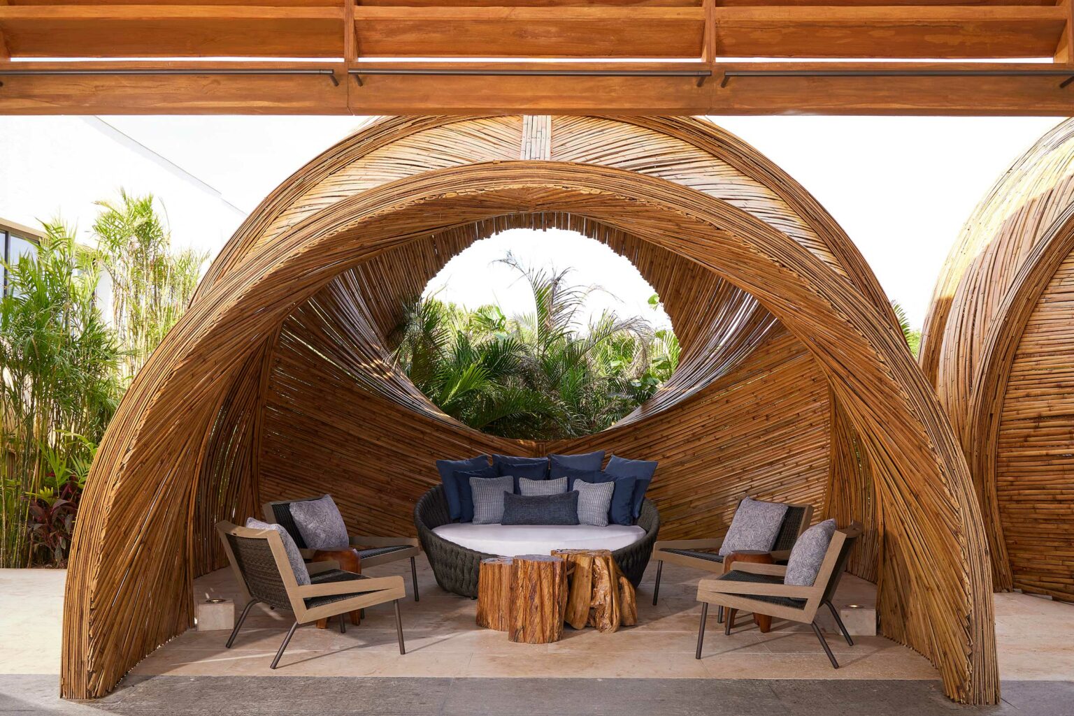 A cozy resort outdoor seating area with a circular wooden canopy, featuring a round cushioned couch with blue pillows and two armchairs. Wooden stump tables add a rustic touch, surrounded by lush greenery. The atmosphere is serene and inviting.