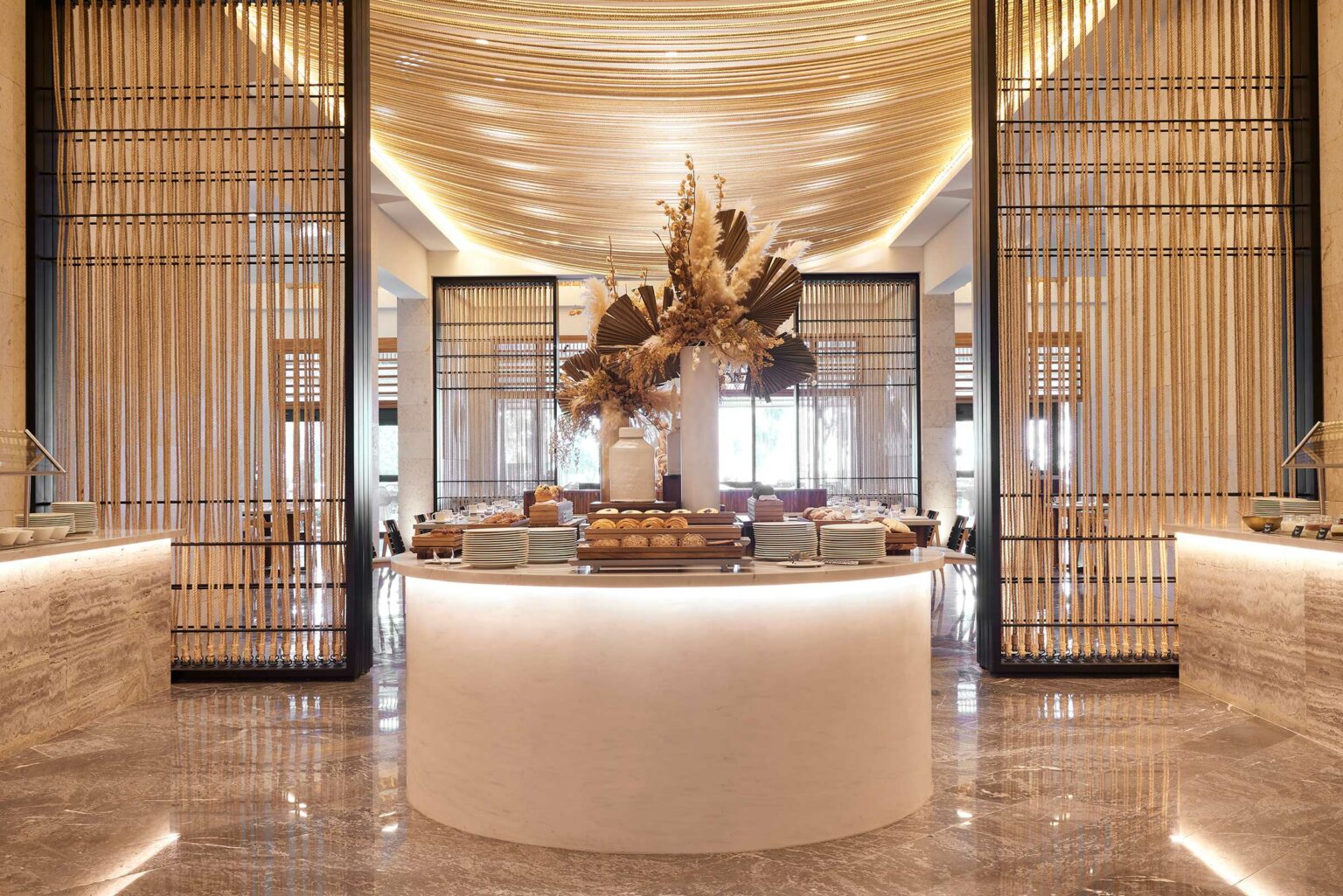 Sophisticated resort all day dining entrance with a round marble counter, dried floral centerpiece, ambient lighting, and elegant pattern screens, conveying luxury.