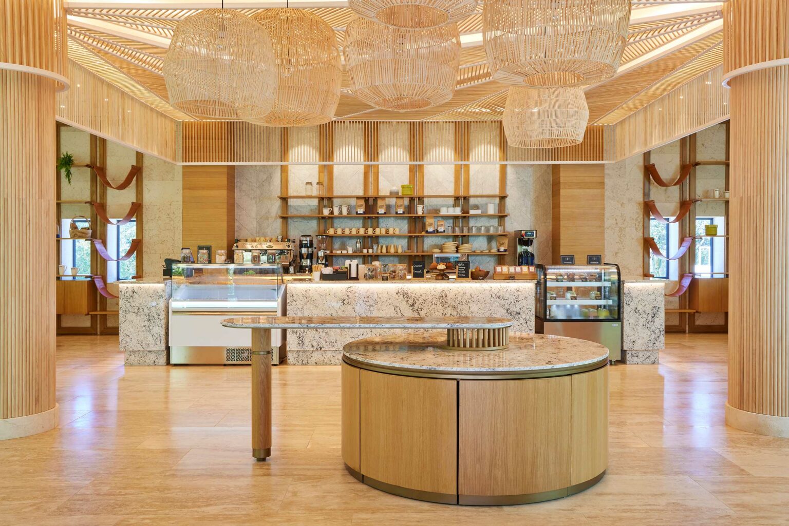 Modern resort café area with wooden accents and marble countertops. Shelves display mugs and coffee products. Large wicker lights hang overhead, creating a cozy atmosphere.
