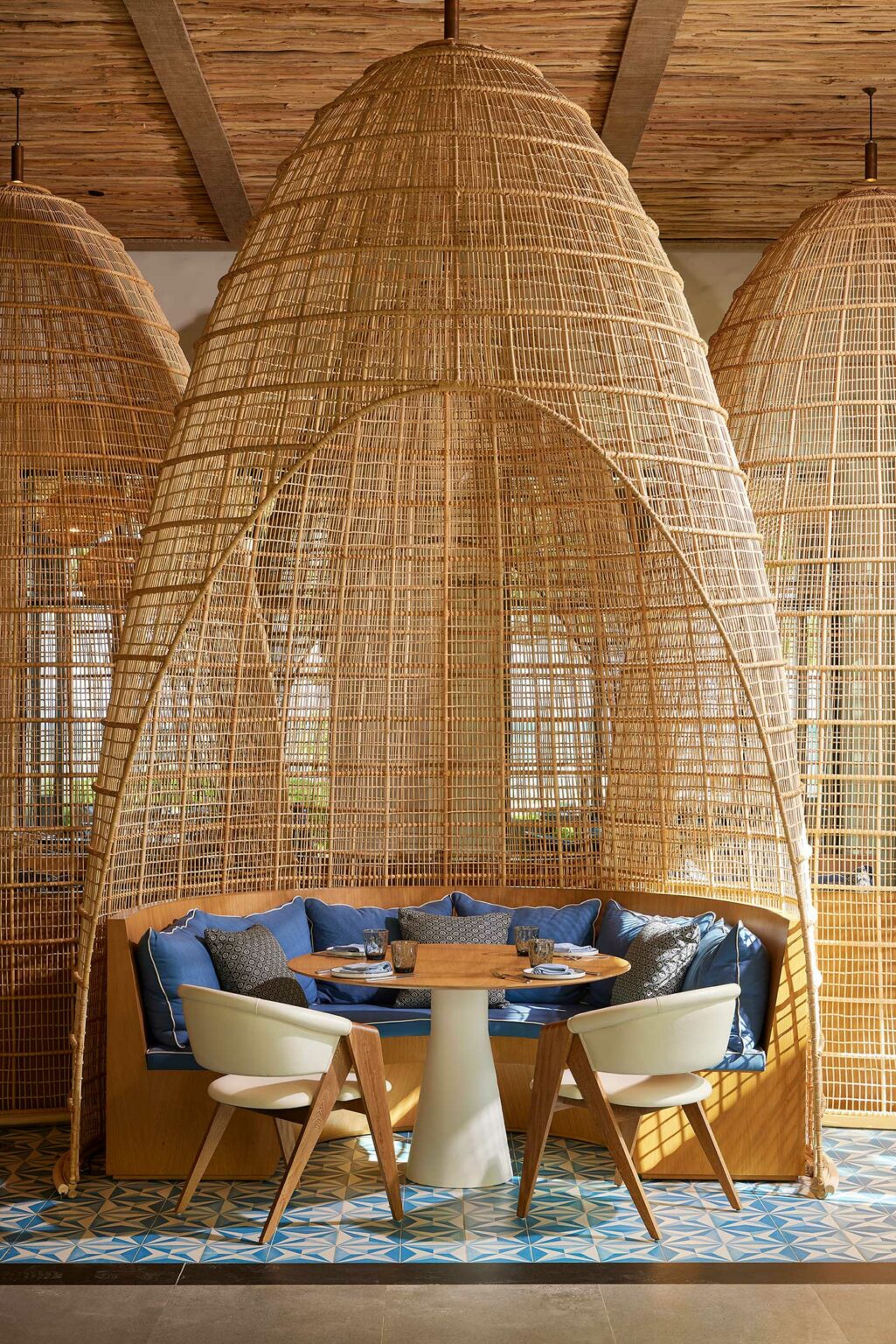 Stylish resort dining area with a round table, blue cushions, and white chairs under a tall, woven canopy. The space is cozy with a serene ambiance.