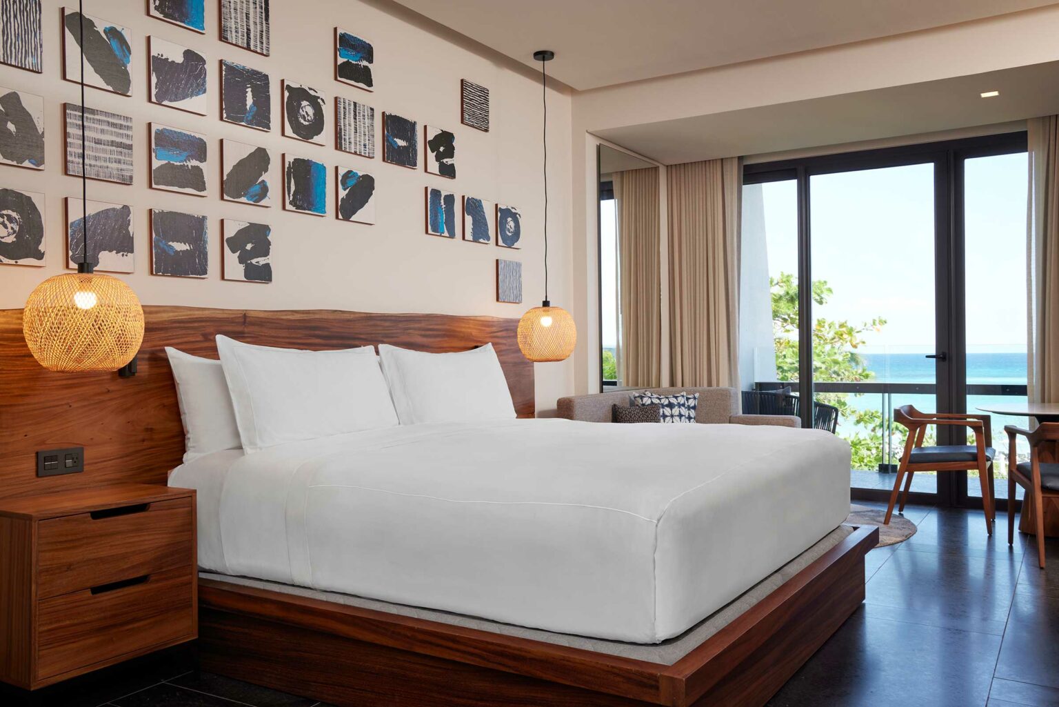 A serene bedroom with a large wooden bed, white bedding, and artistic wall decor. Warm pendant lights hang above. Glass doors reveal an ocean view.