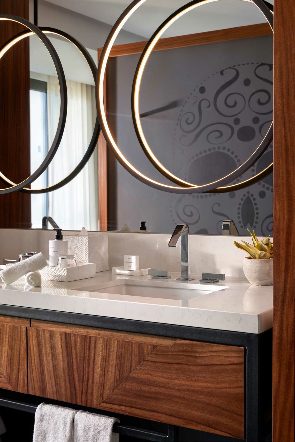 Modern suite bathroom with a white countertop and wooden cabinet. Circular mirrors with lights reflect a patterned gray wall. Fresh towel and plant add elegance.