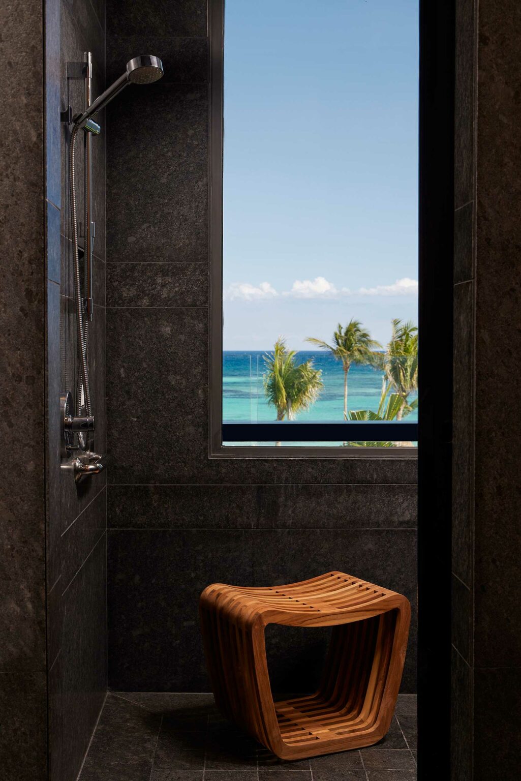 Dark gray shower interior with a wooden stool, featuring a large window displaying a clear blue ocean view and palm trees, creating a tranquil setting.