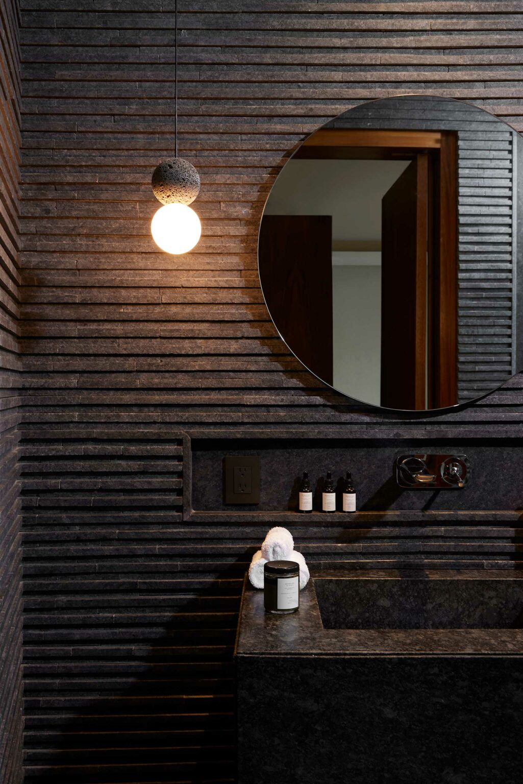 Dimly lit modern suite bathroom with a dark textured wall, round mirror, and simple pendant light. The minimalist sink holds a candle and folded towel.