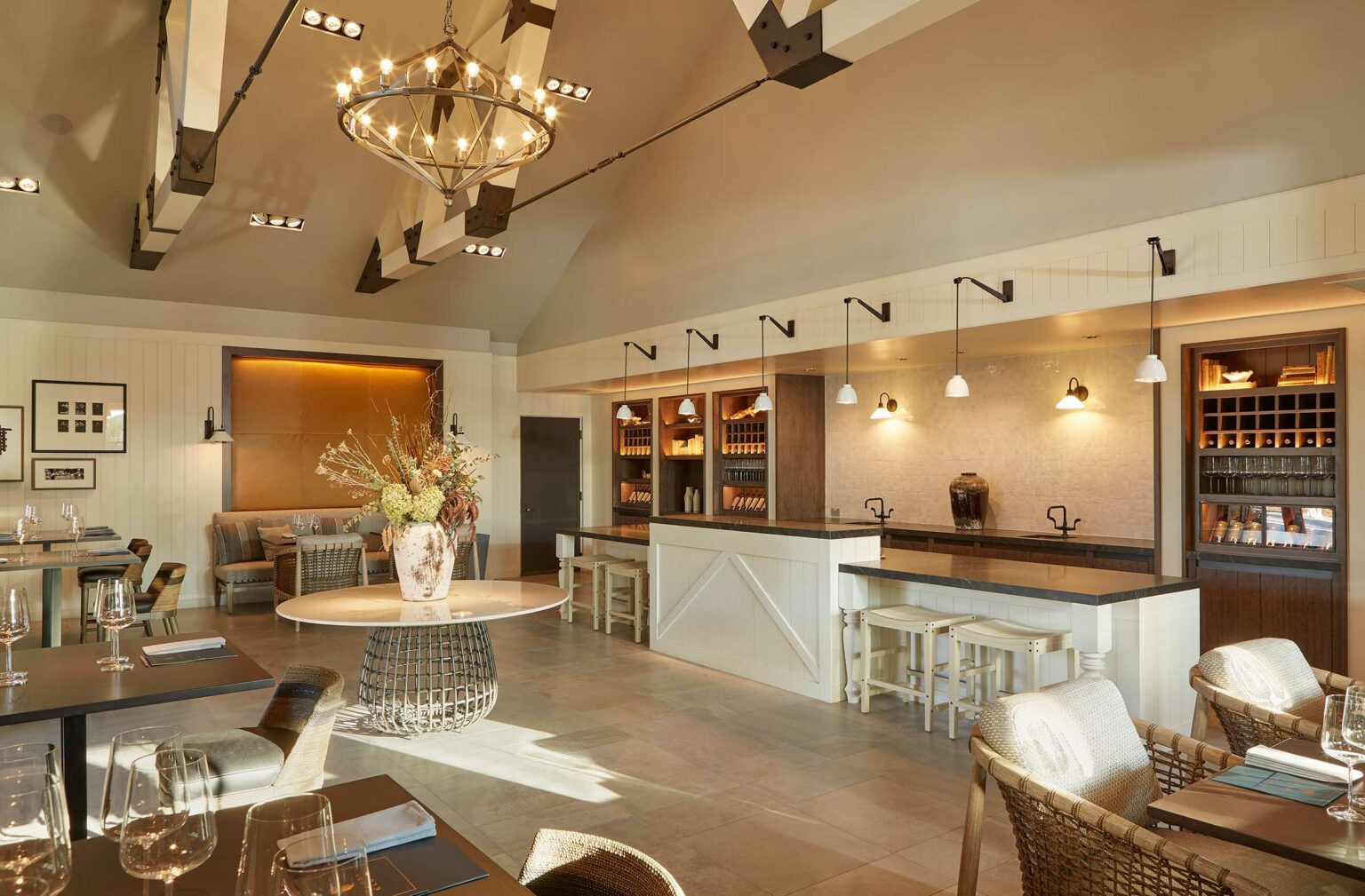 Rustic dining room with a cozy atmosphere set in a wine resort. Features wooden tables, wicker chairs, elegant chandeliers, and a large vase of dried flowers on a central table.