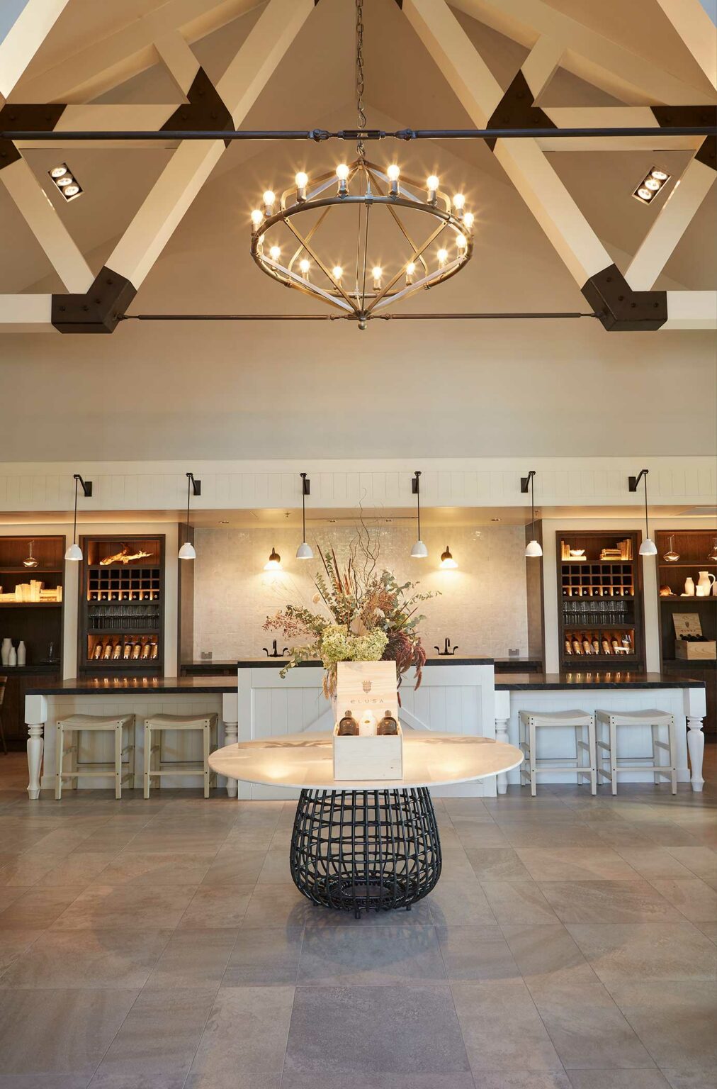 Spacious, elegant interior of a wine resort with wooden beams, circular chandelier, and a central table with flowers. Background: bar area with stools and wall lights.