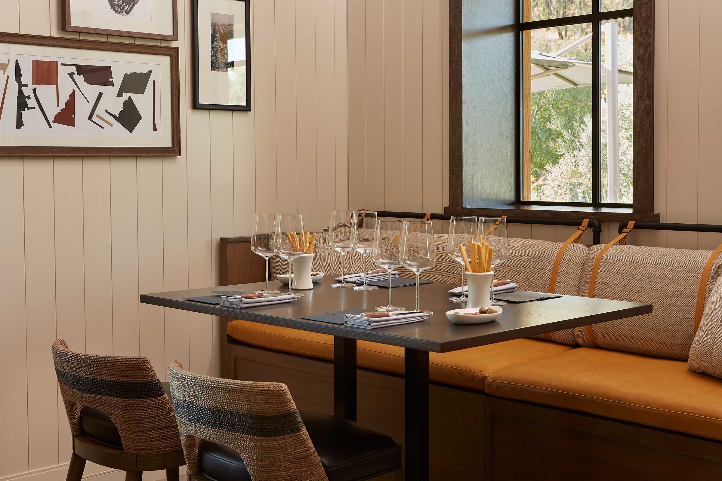 Cozy dining corner of a wine resort with a black table set for four. Glasses and cutlery arranged neatly. Soft beige and orange tones with modern art on walls.