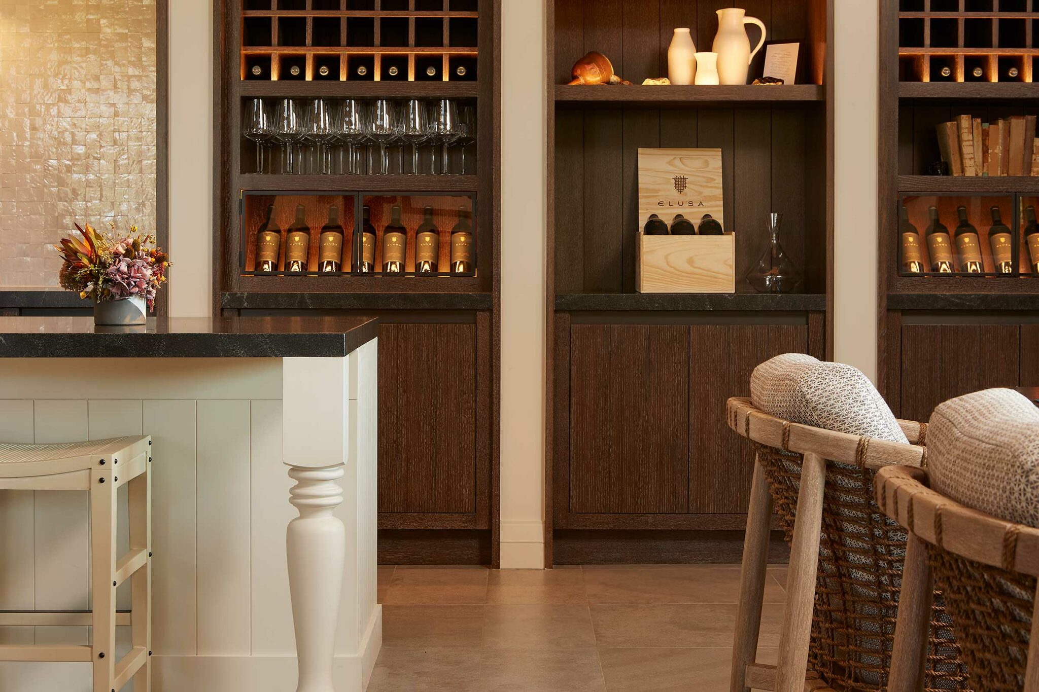 A cozy wine bar corner of a wine resort with wooden shelves displaying wine bottles and glasses. Elegant bar counter with a flower vase. Warm, inviting ambiance.