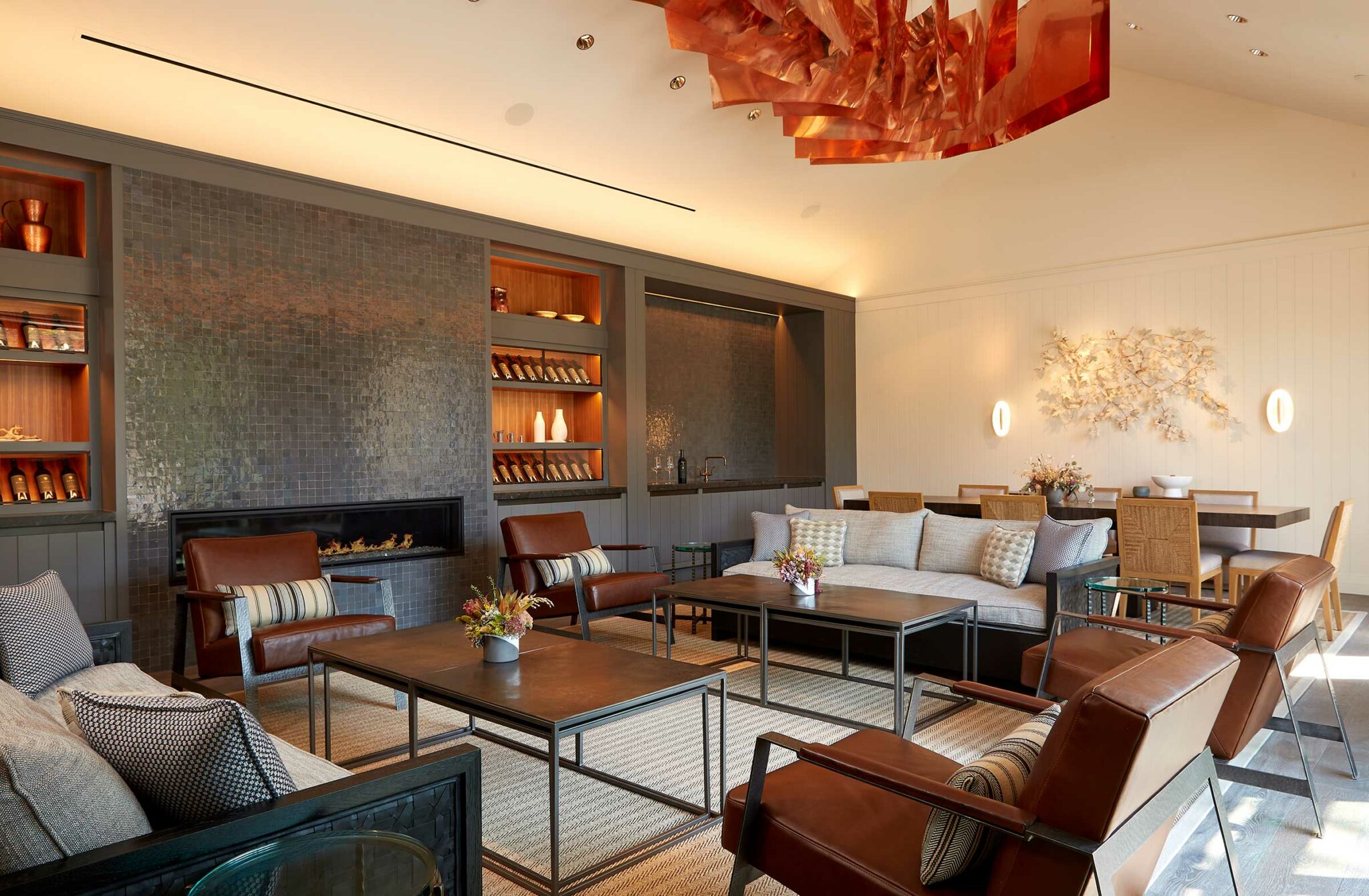 Stylish lounge of a winery resort with leather chairs, a modern sofa, and a central table under an abstract orange chandelier. Warm lighting and art accents create a cozy ambiance.
